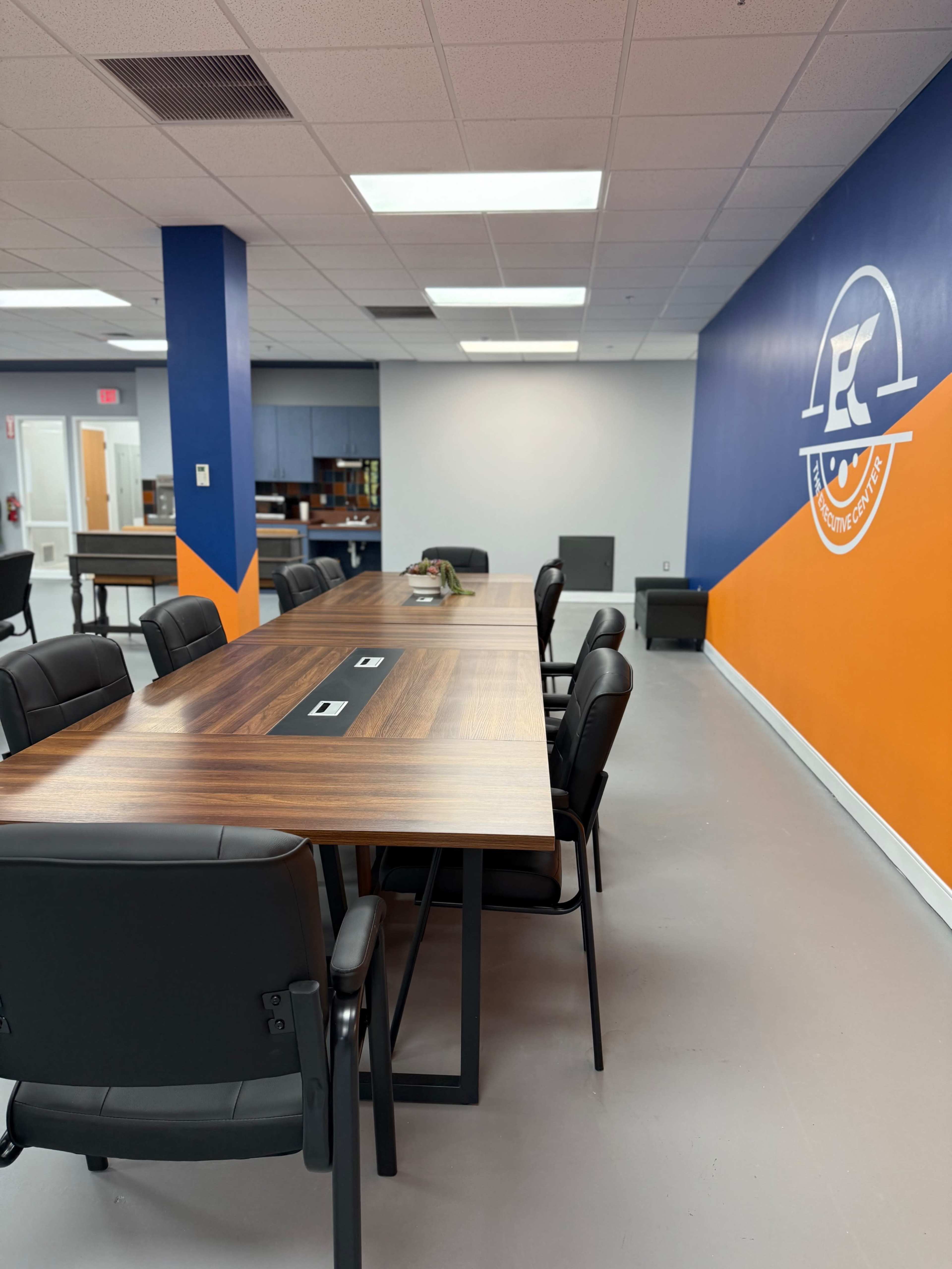 The image shows a modern conference room with a long wooden table surrounded by black chairs, featuring an orange and blue wall with a logo.
