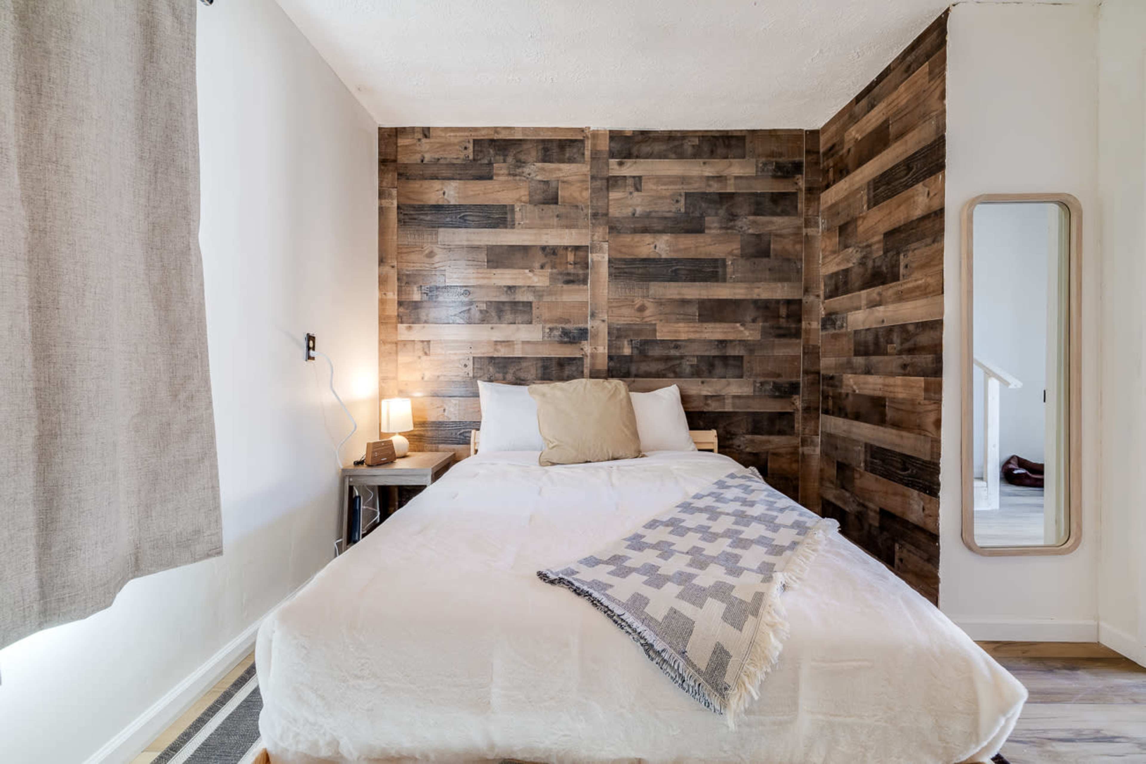 A neatly arranged bedroom features a wooden accent wall, a double bed with a light blanket, a small lamp, and a full-length mirror.