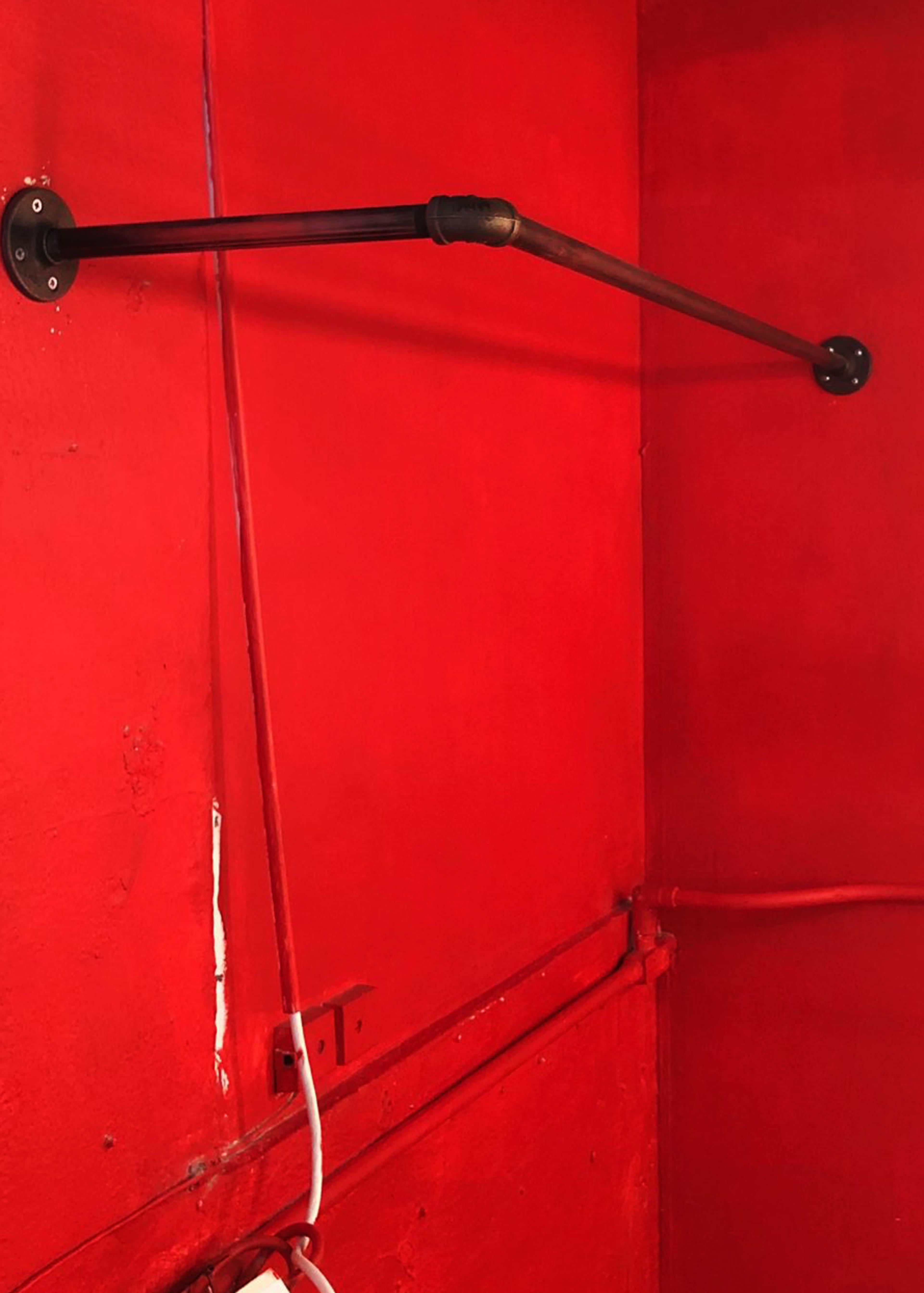 A black metal rod is mounted on a red wall with visible pipes and electrical wiring nearby.