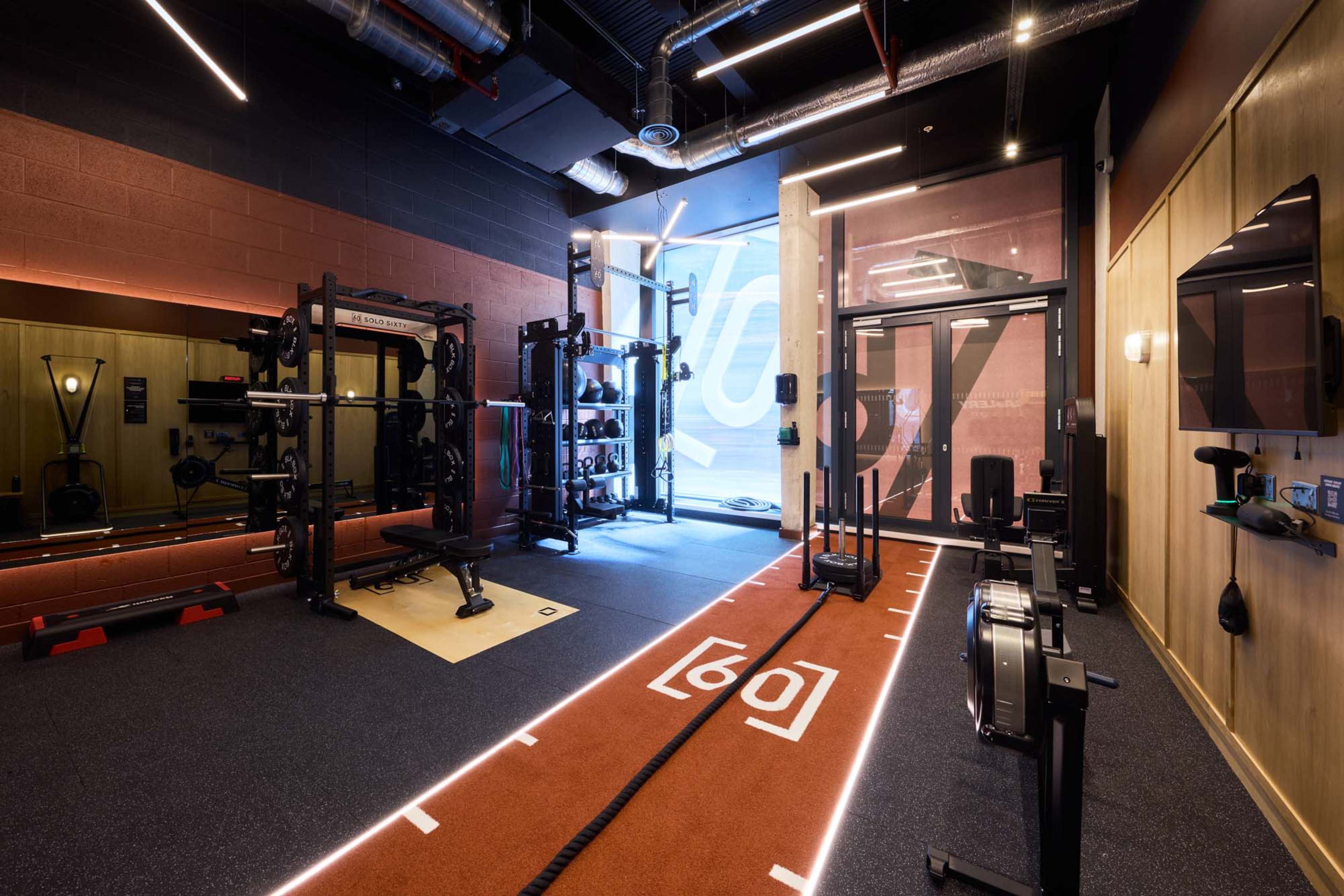 The image shows a modern gym equipped with various fitness equipment, including a rowing machine, weights, and exercise mats, along a brightly lit area with a turf track.