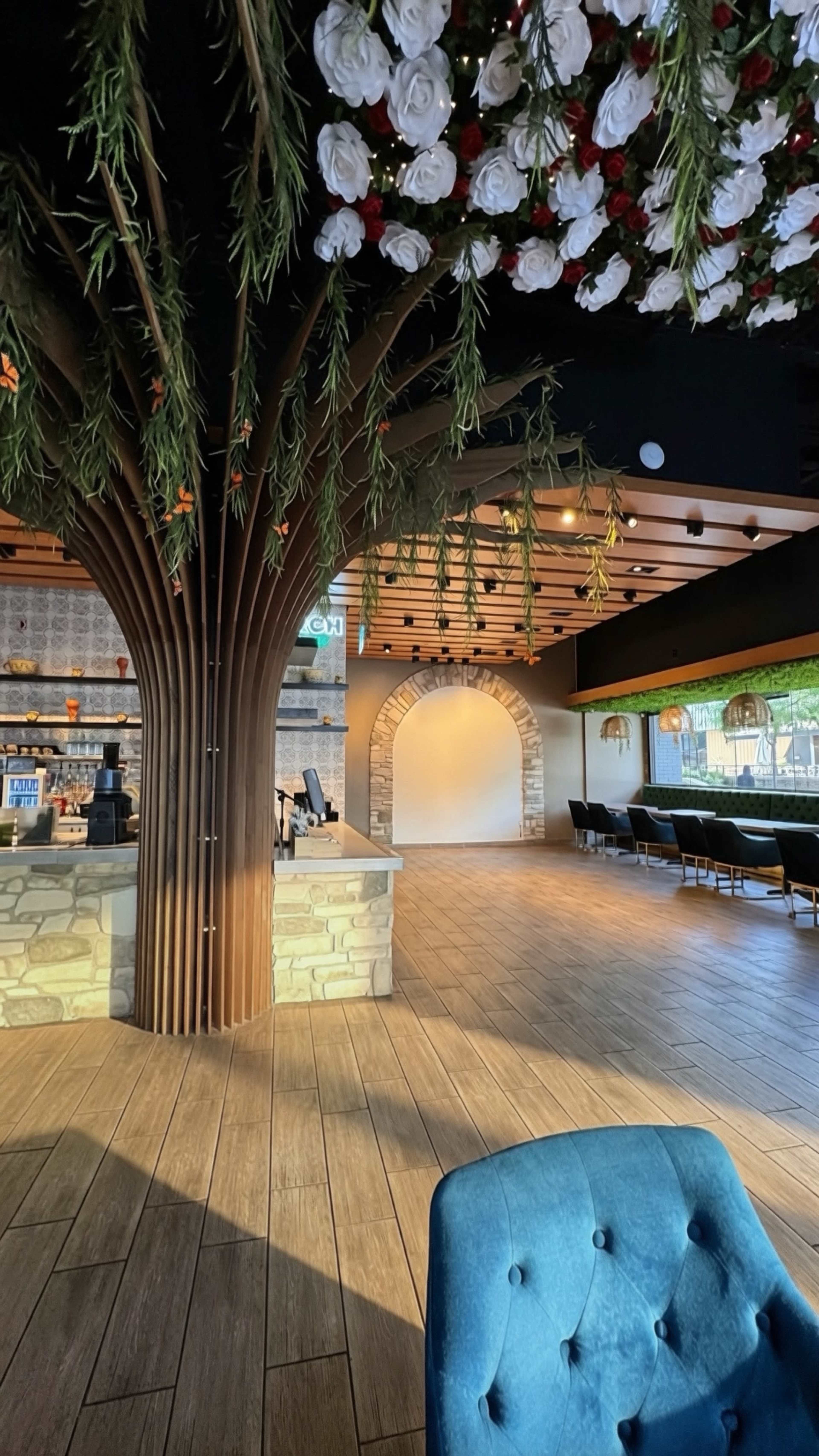 The image depicts a modern interior space featuring a bar area with stone accents under a large, decorative tree structure adorned with flowers, and an empty archway in the background.