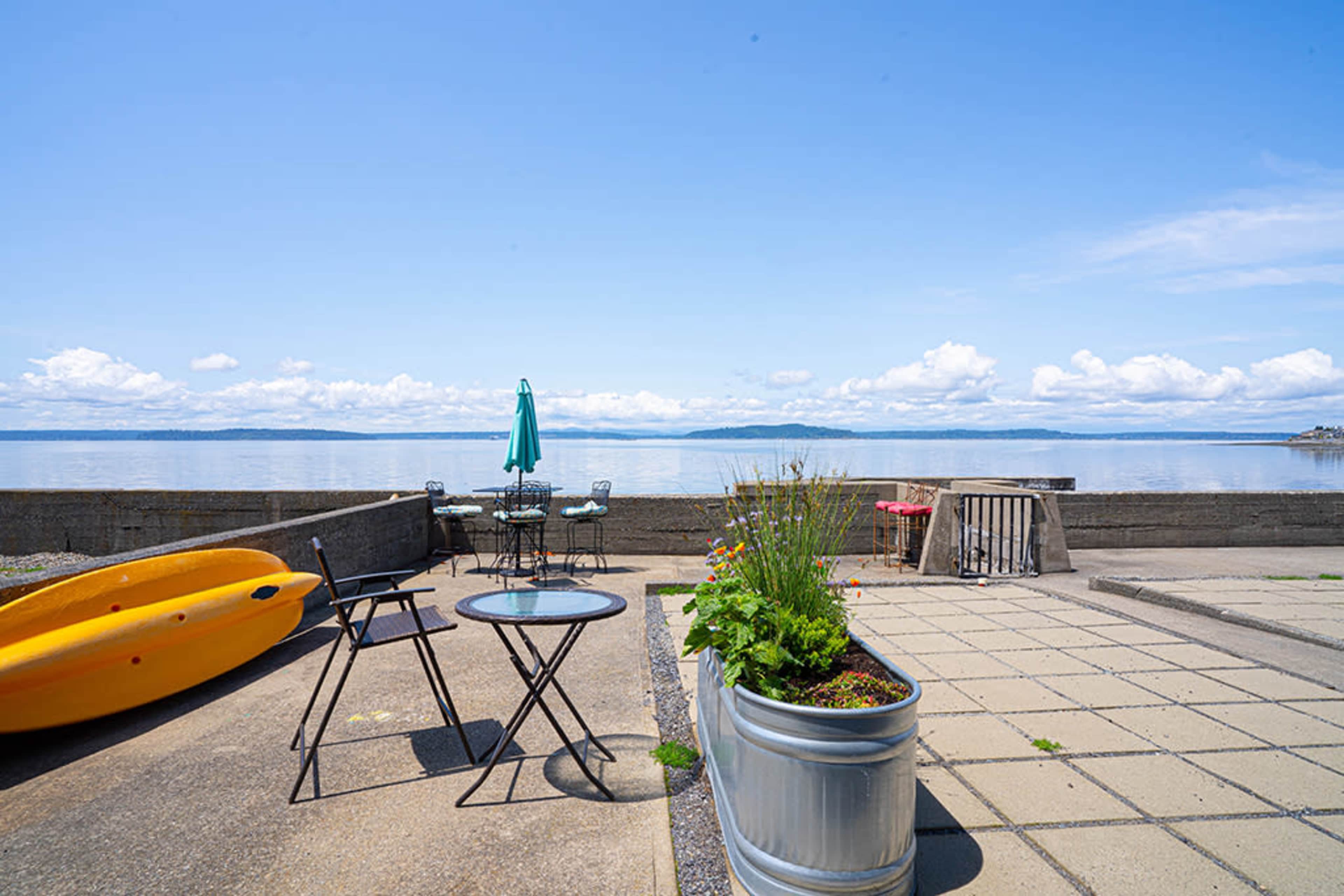 A kayak and patio furniture are arranged on a concrete area overlooking a calm waterfront with distant hills.