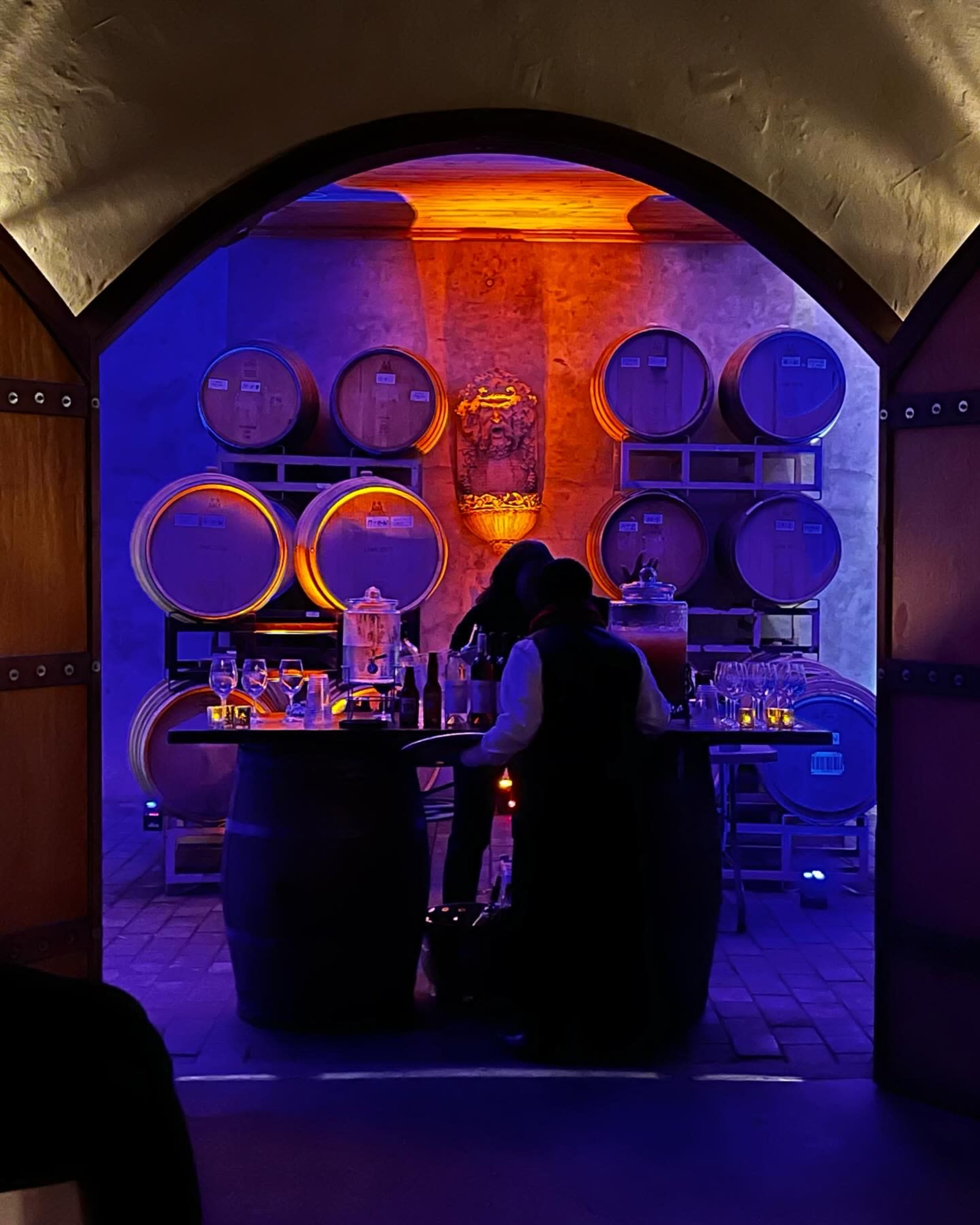 A dimly lit wine cellar features stacked barrels in the background and a bar setup with glasses and bottles in the foreground.
