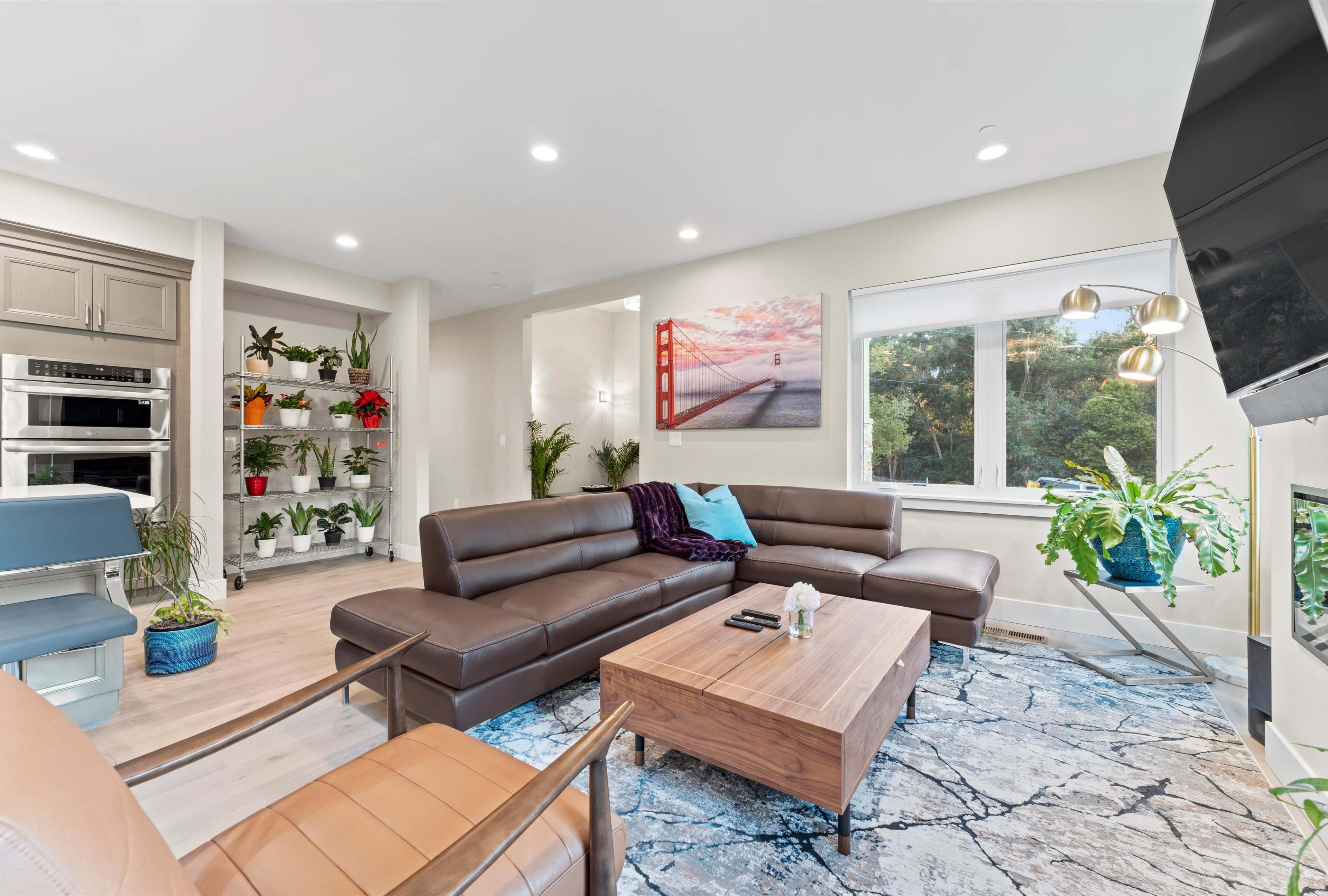 A modern living room with a sectional couch, a wooden coffee table, a large window featuring greenery outside, and decorative plants on shelves.