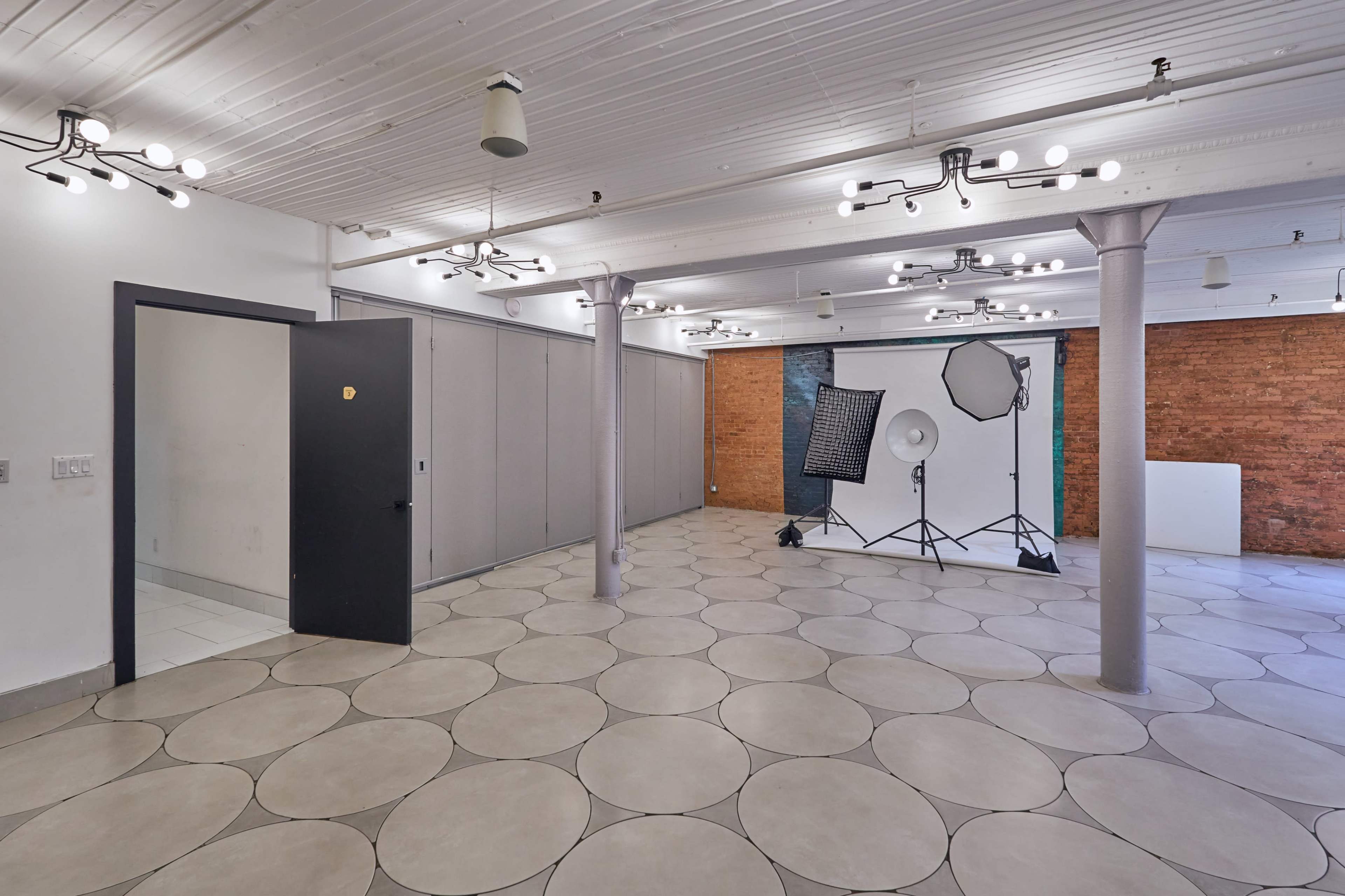 A spacious, well-lit room with circular tile flooring, a black door, and a photography setup featuring softboxes and a seamless backdrop against a brick wall.