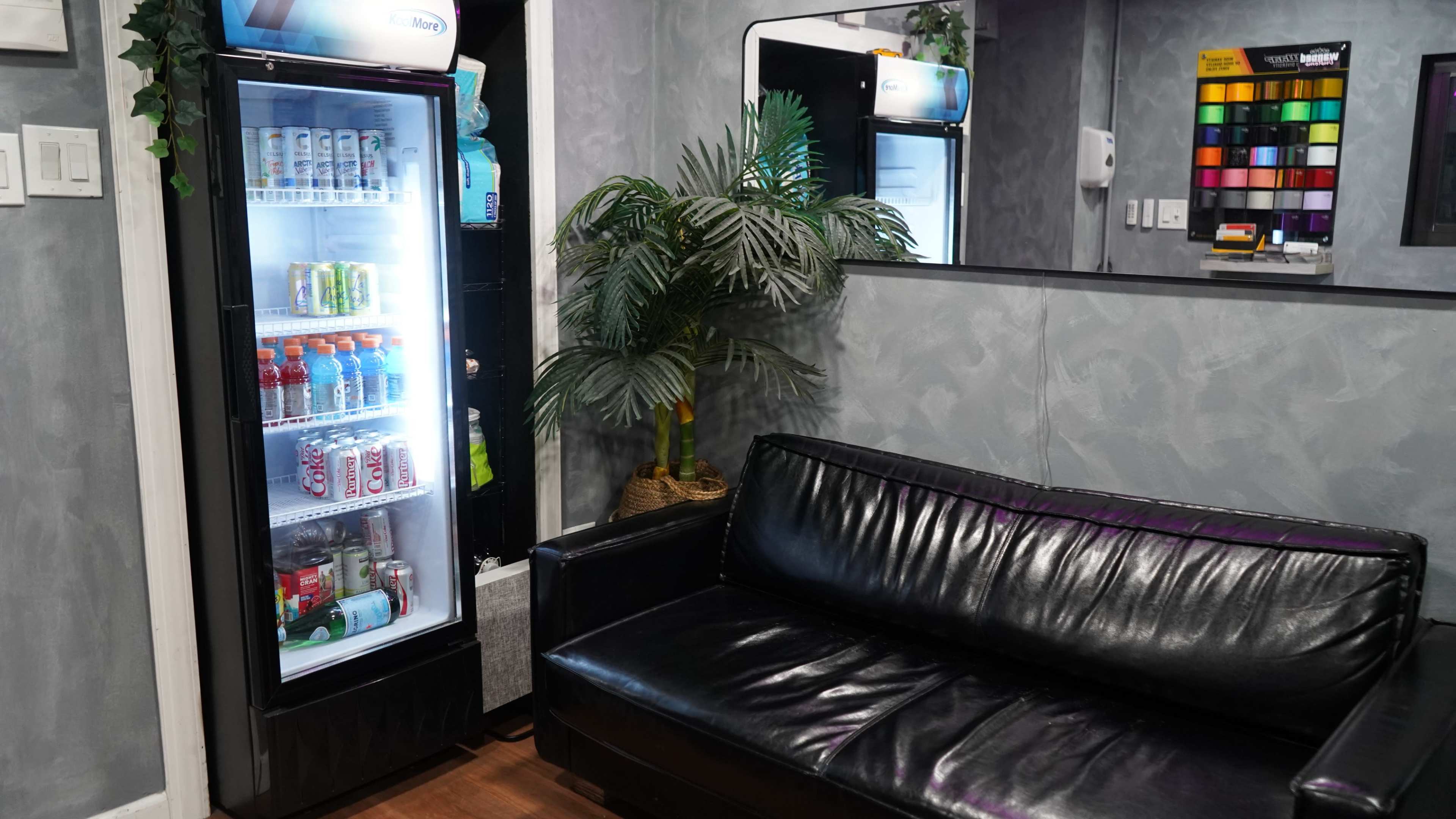 The image shows a small waiting area with a black leather couch next to a refrigerator stocked with beverages and a decorative plant.