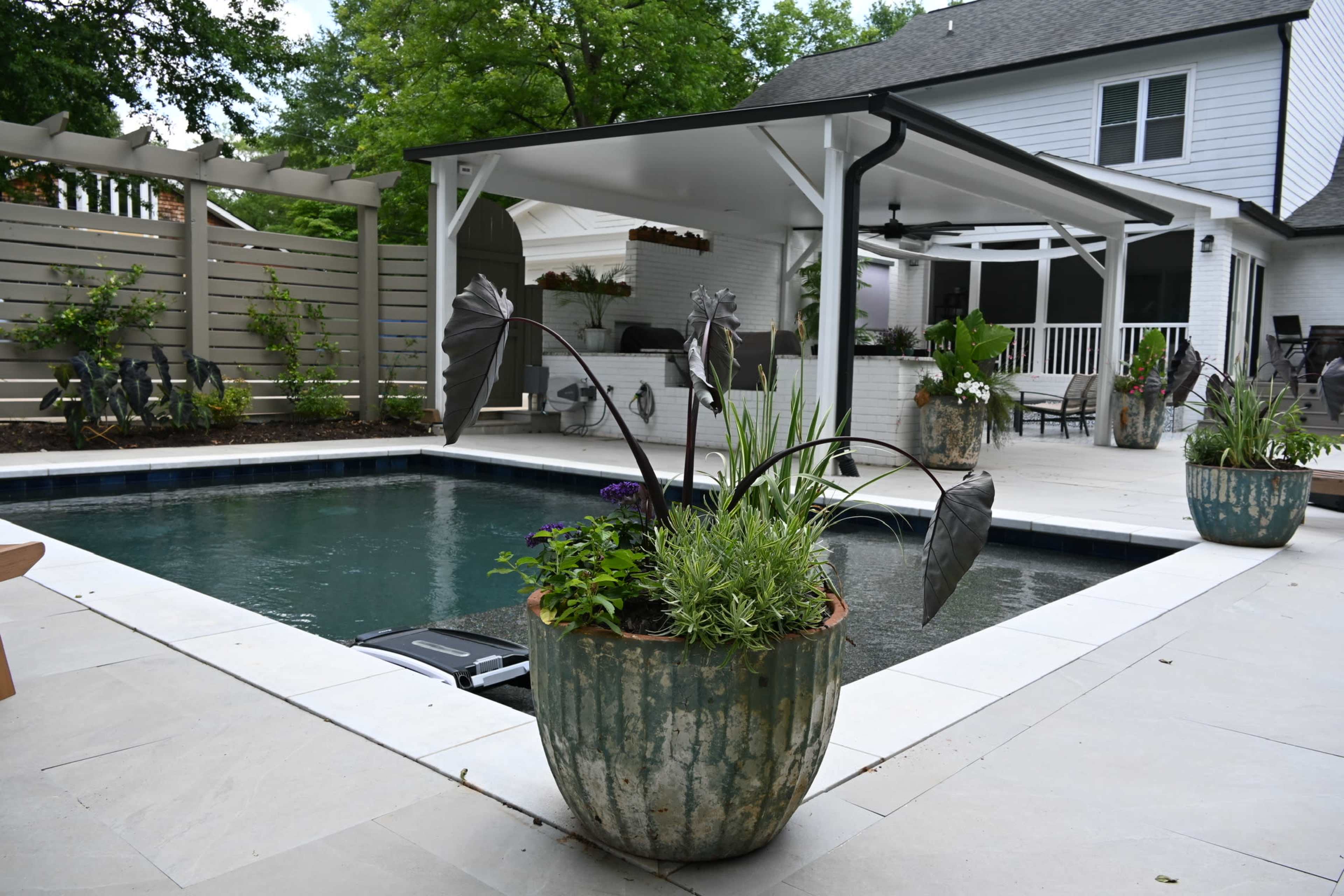 A rectangular swimming pool surrounded by stone tiles and large planters adorned with plants, next to a covered patio area.