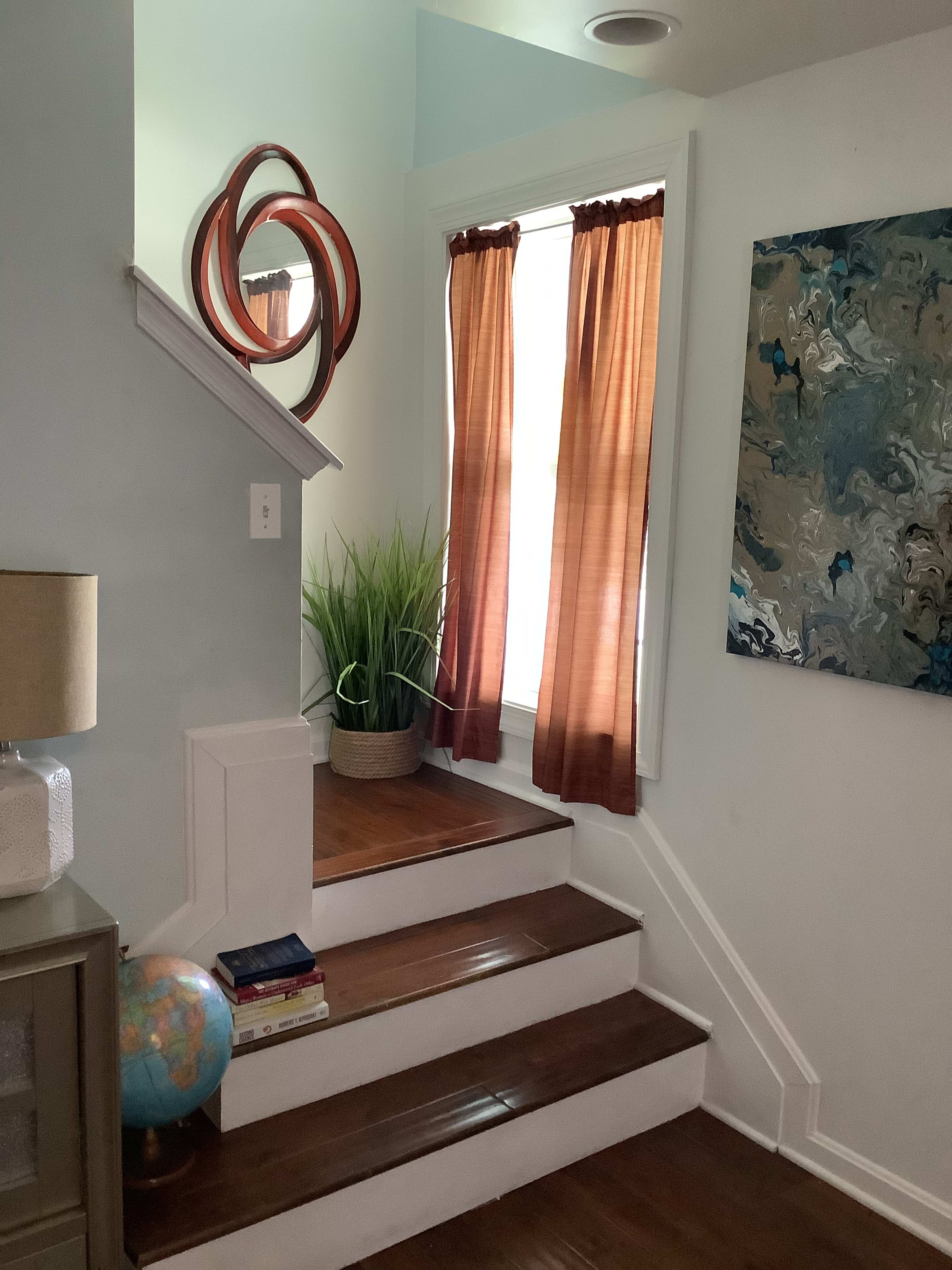 A staircase with wooden steps leads up to a window draped with orange curtains, accompanied by a large decorative mirror and a plant in a pot.