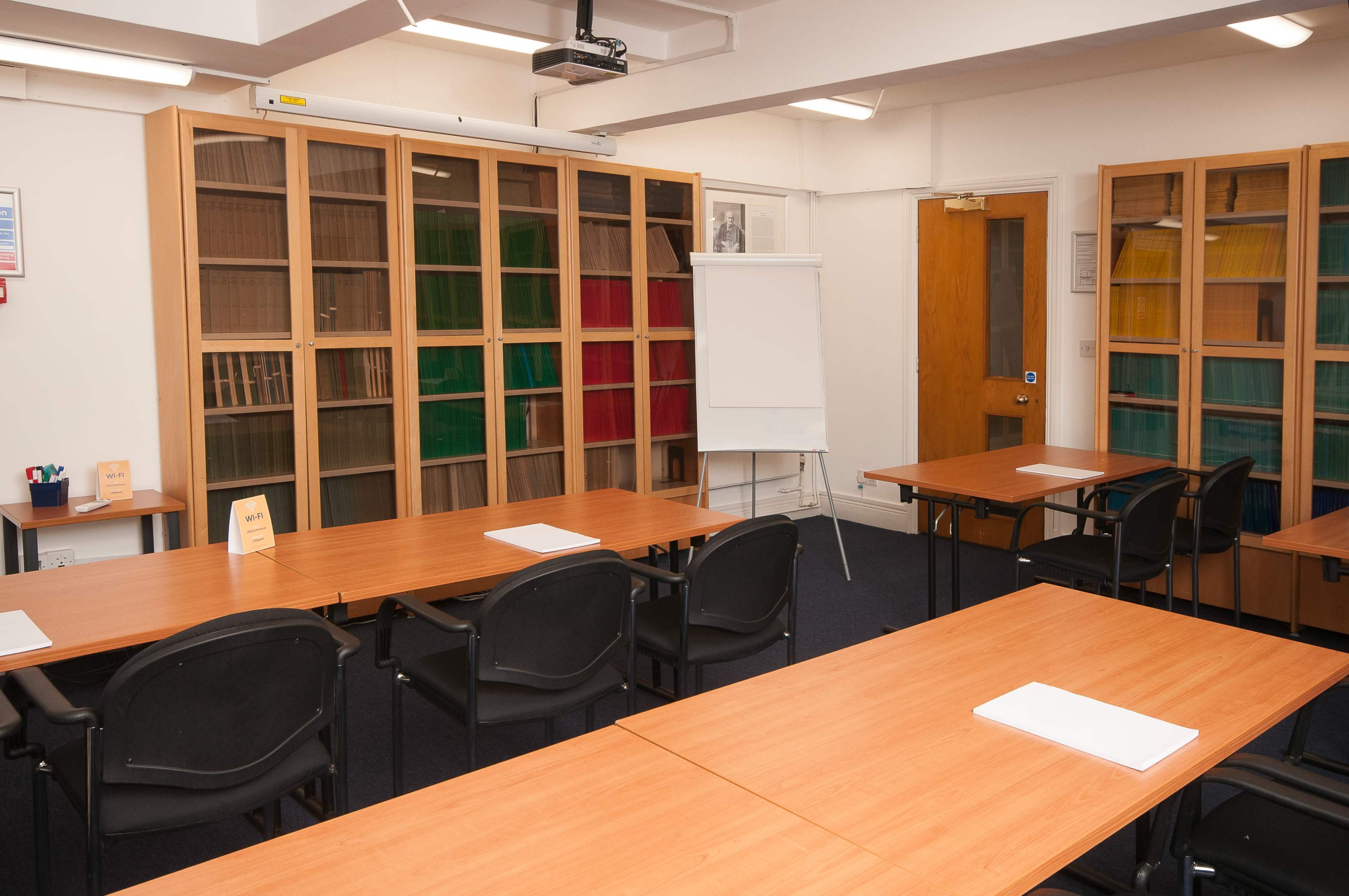A classroom setup features several tables and chairs arranged for a meeting, with a blank flip chart and shelves containing colored files in the background.