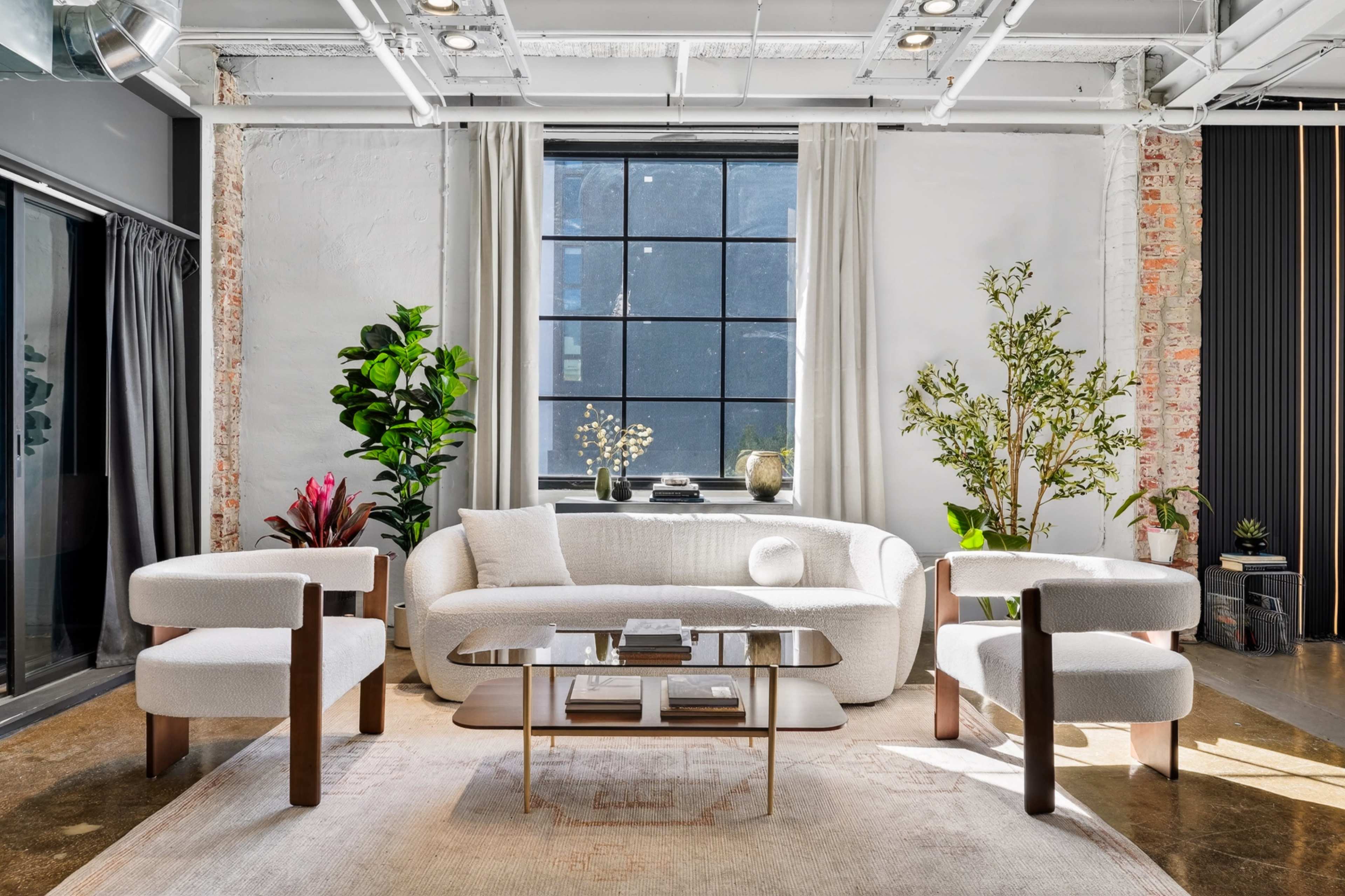 The image shows a stylish living room featuring a light-colored sofa, two modern armchairs, a glass coffee table, and several indoor plants near large windows.