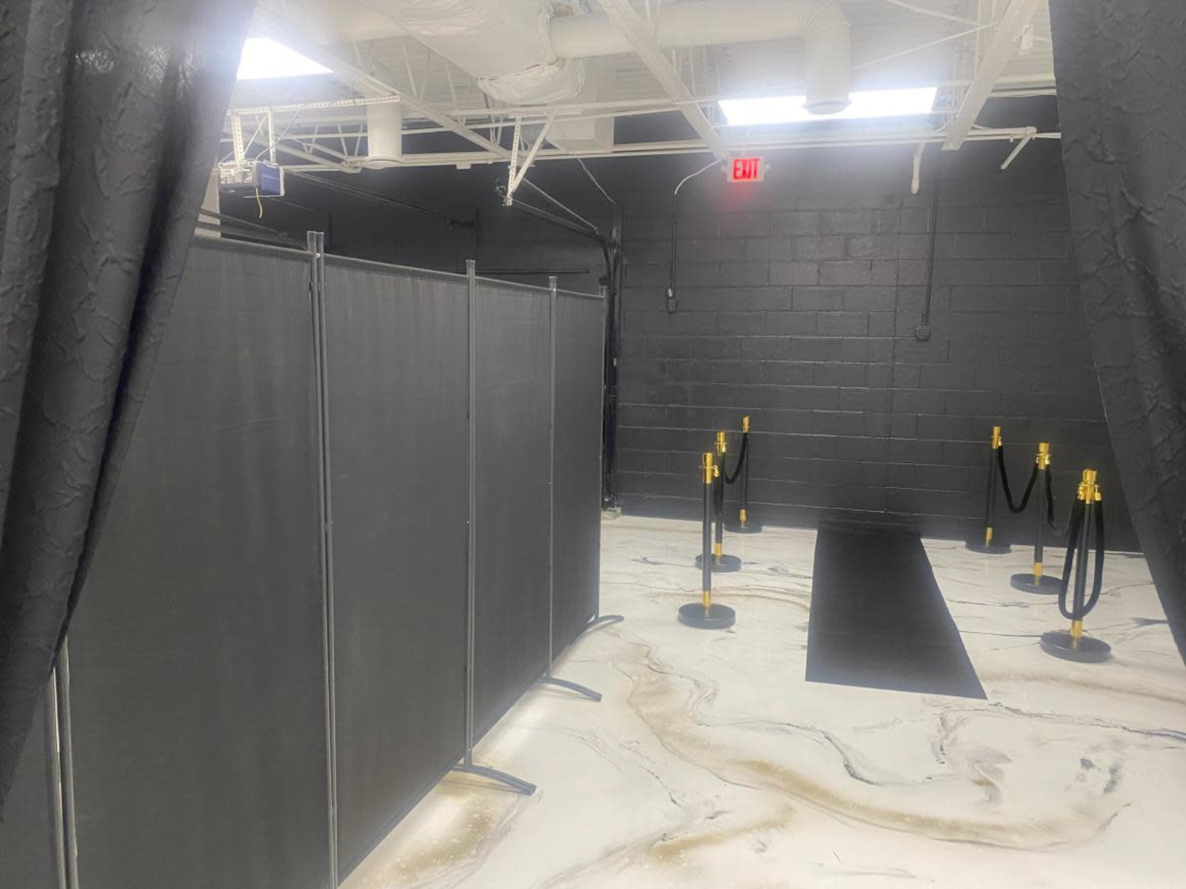 The image shows a dimly lit hallway with black curtains, partitions, and red exit lights leading to a carpeted area with stanchions.