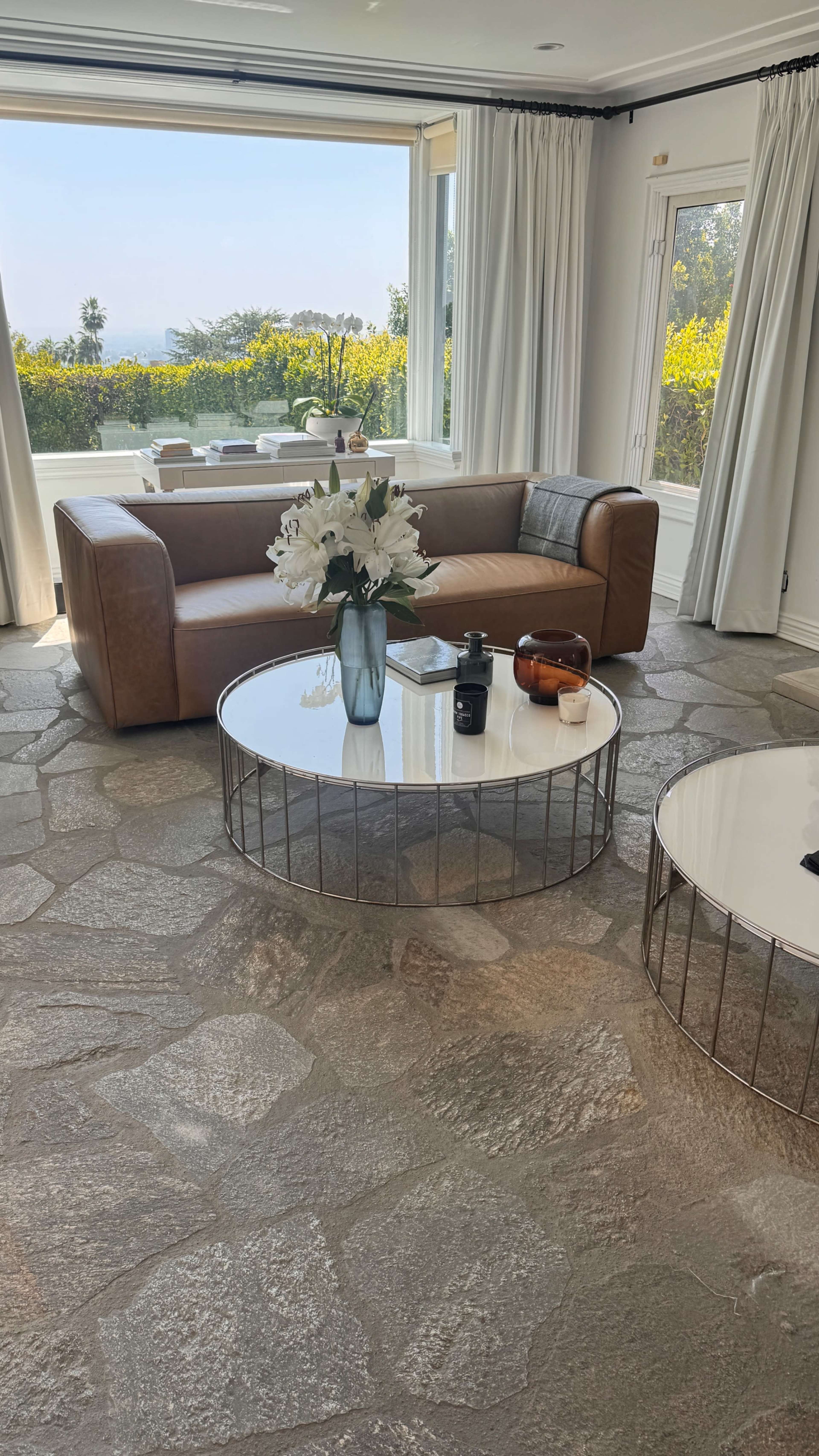 A leather sofa is positioned in a bright room with large windows, showcasing a view of greenery, and two round coffee tables sit on a stone floor.