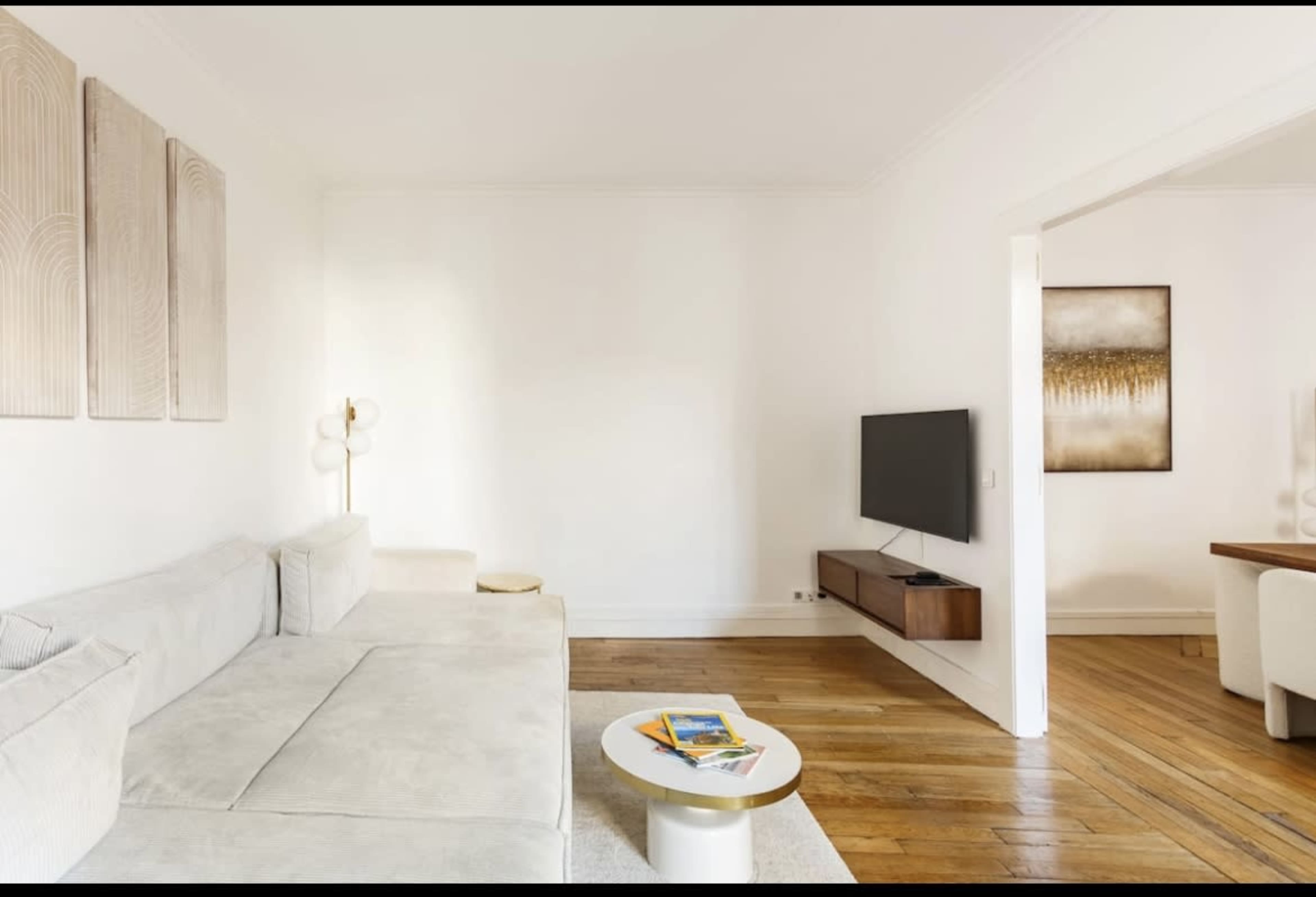 A modern living room with a light-colored sofa, a small circular coffee table, a mounted television on the wall, and a wooden dining table visible in the adjacent space.