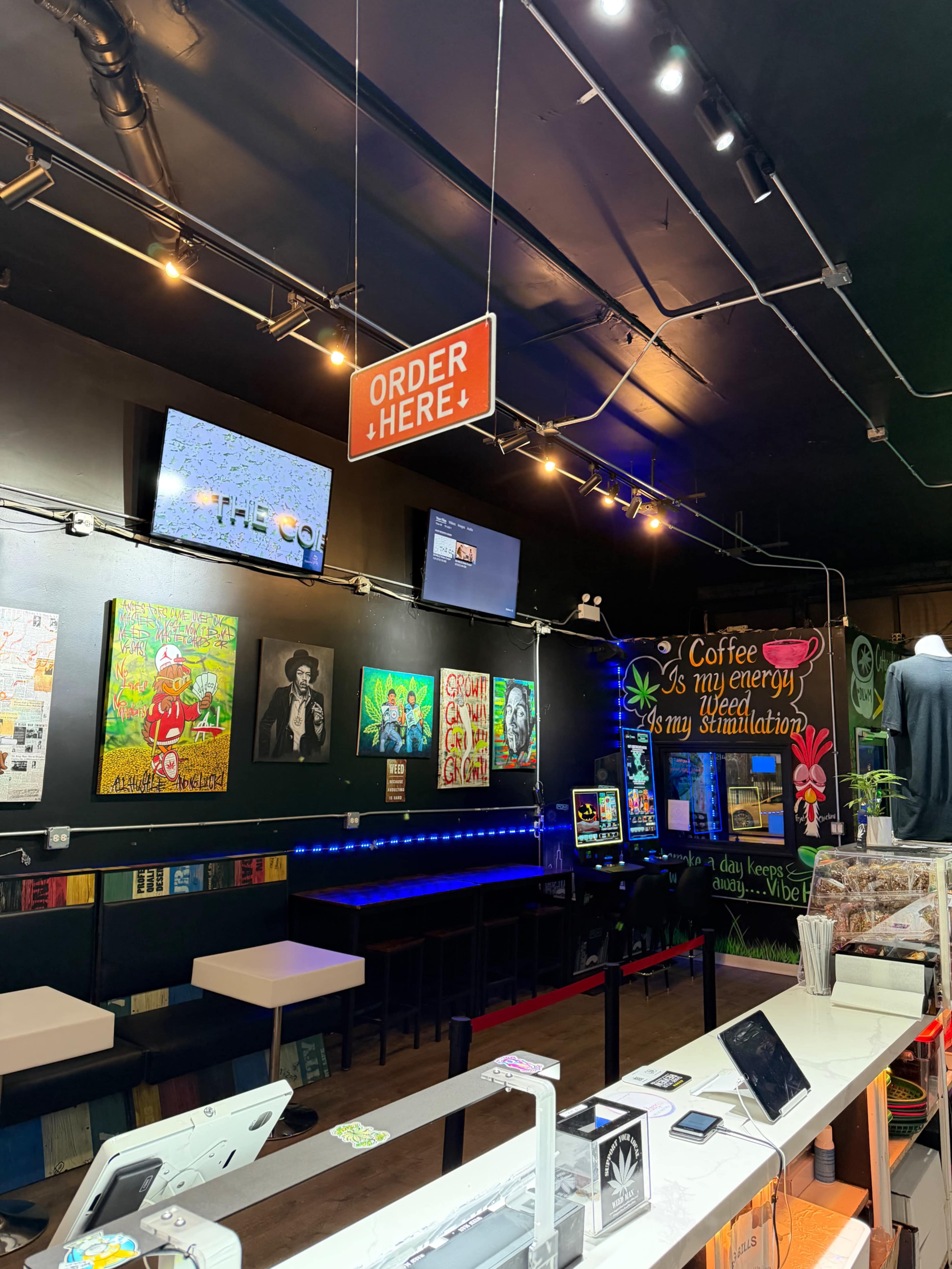 The image shows an ordering area in a colorful café with wall art, multiple screens, and seating.
