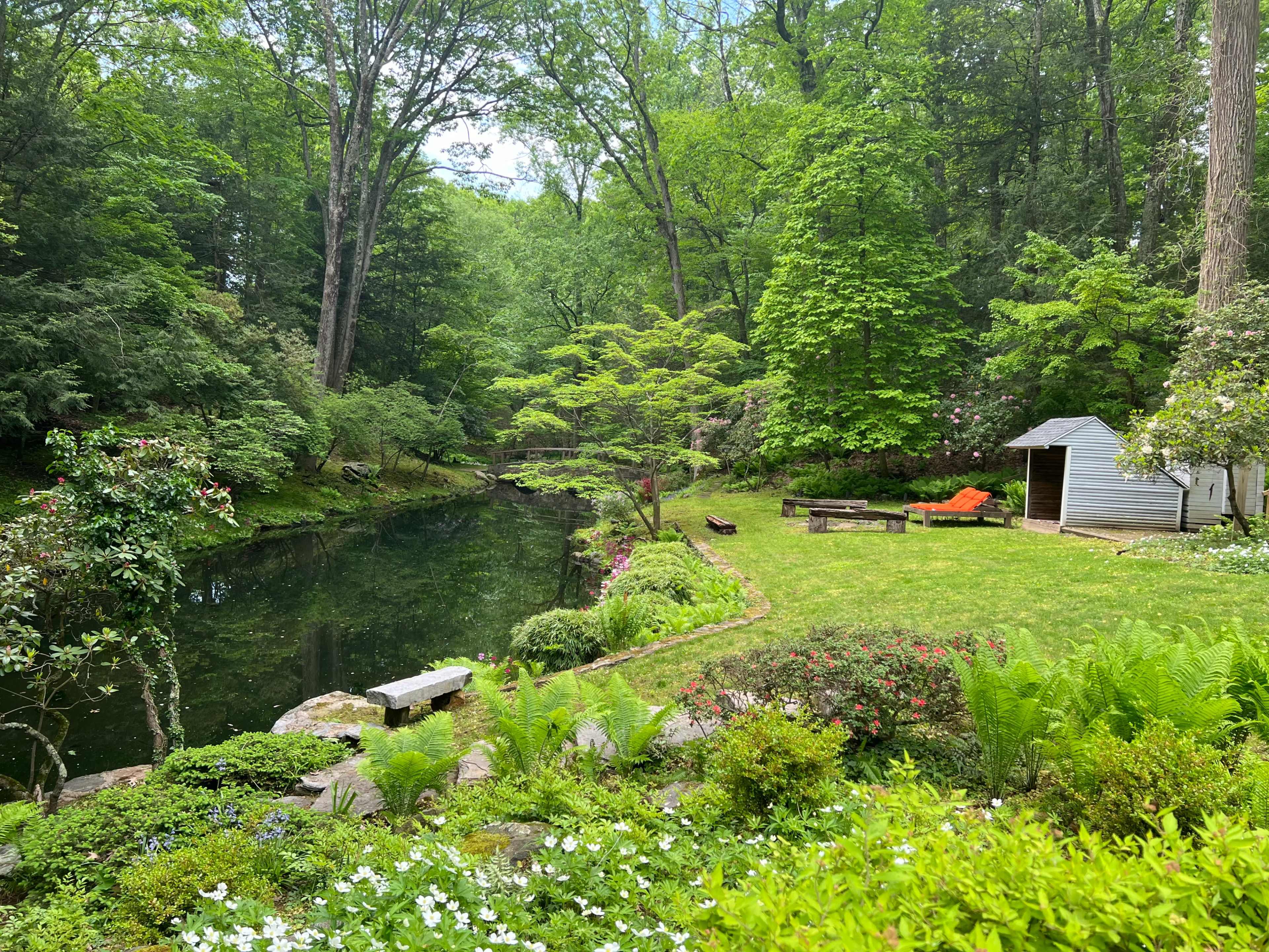 A serene garden with a pond surrounded by lush greenery, a small white shed, and seating areas.