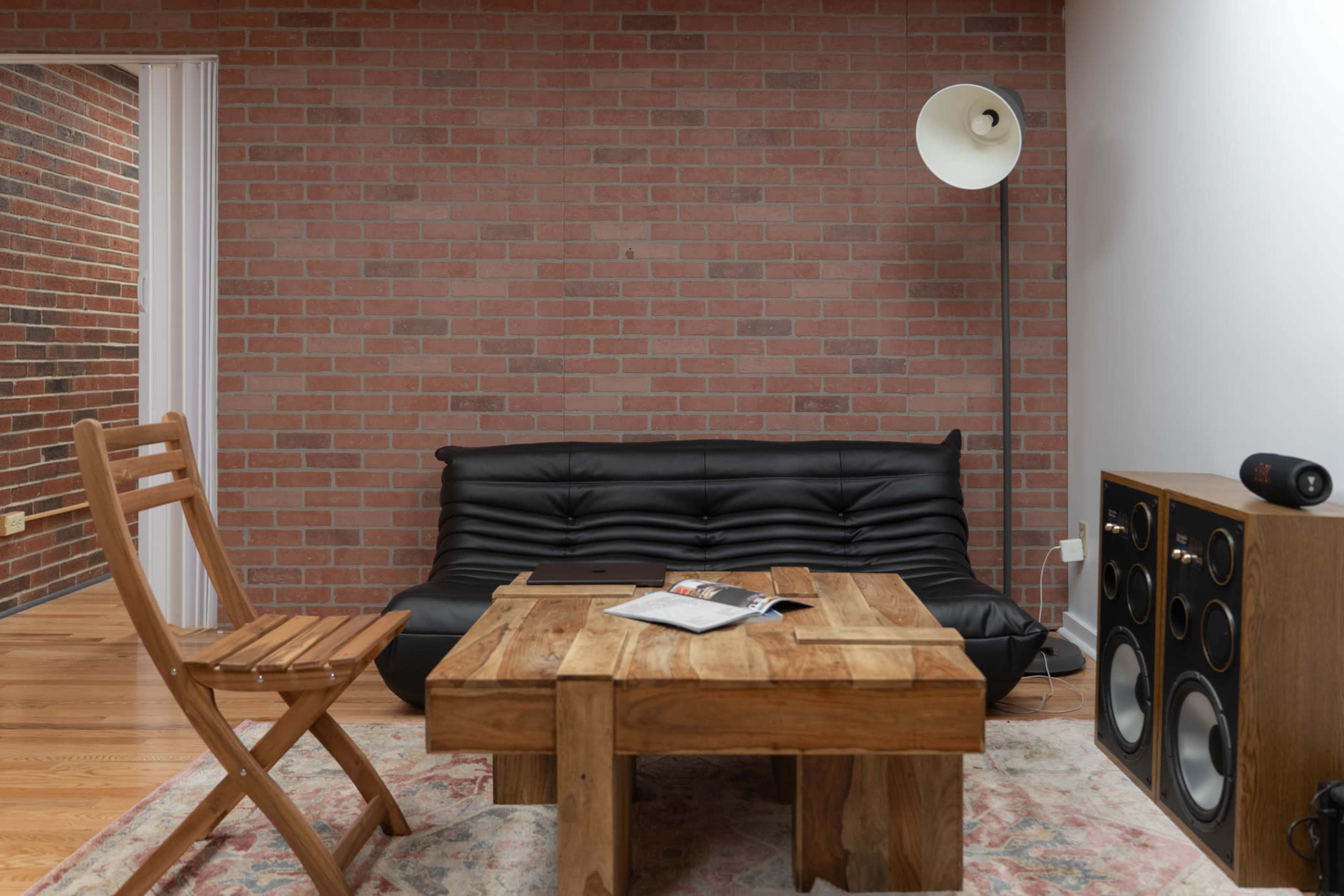 The image depicts a cozy living area featuring a black sofa, a wooden coffee table, a floor lamp, and speakers against a brick wall.