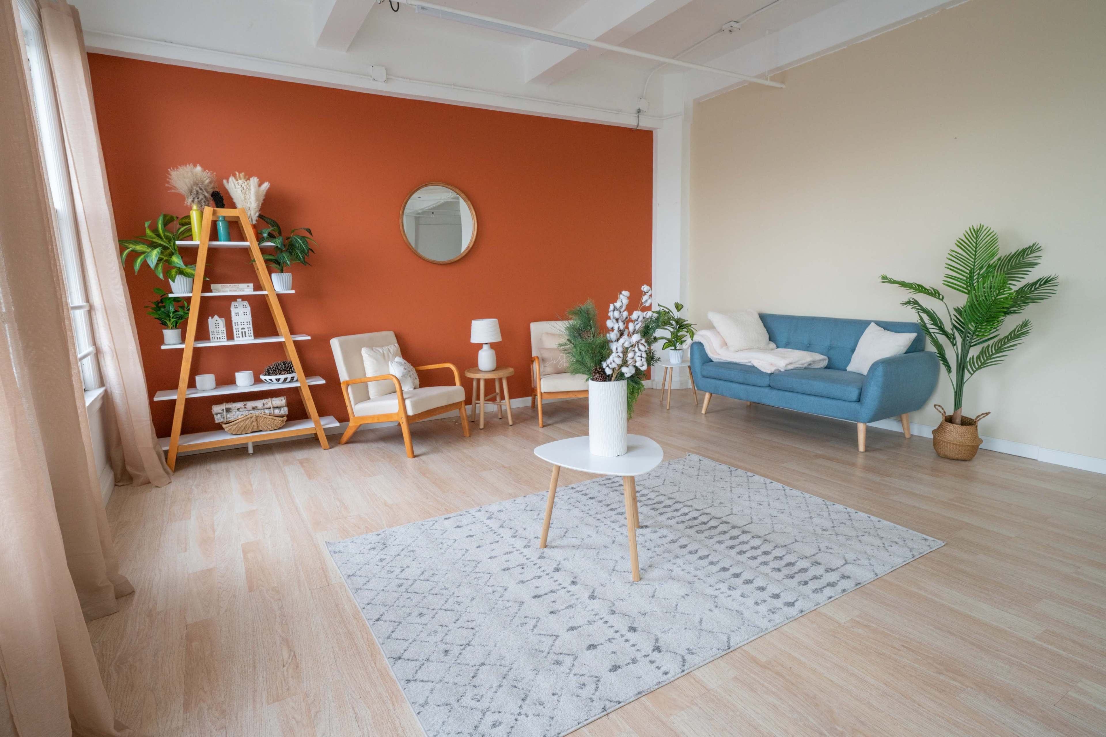 The image shows a modern, well-lit living room with an orange accent wall, a blue sofa, a wooden ladder shelf with decorative items, and a white circular coffee table on a patterned rug.