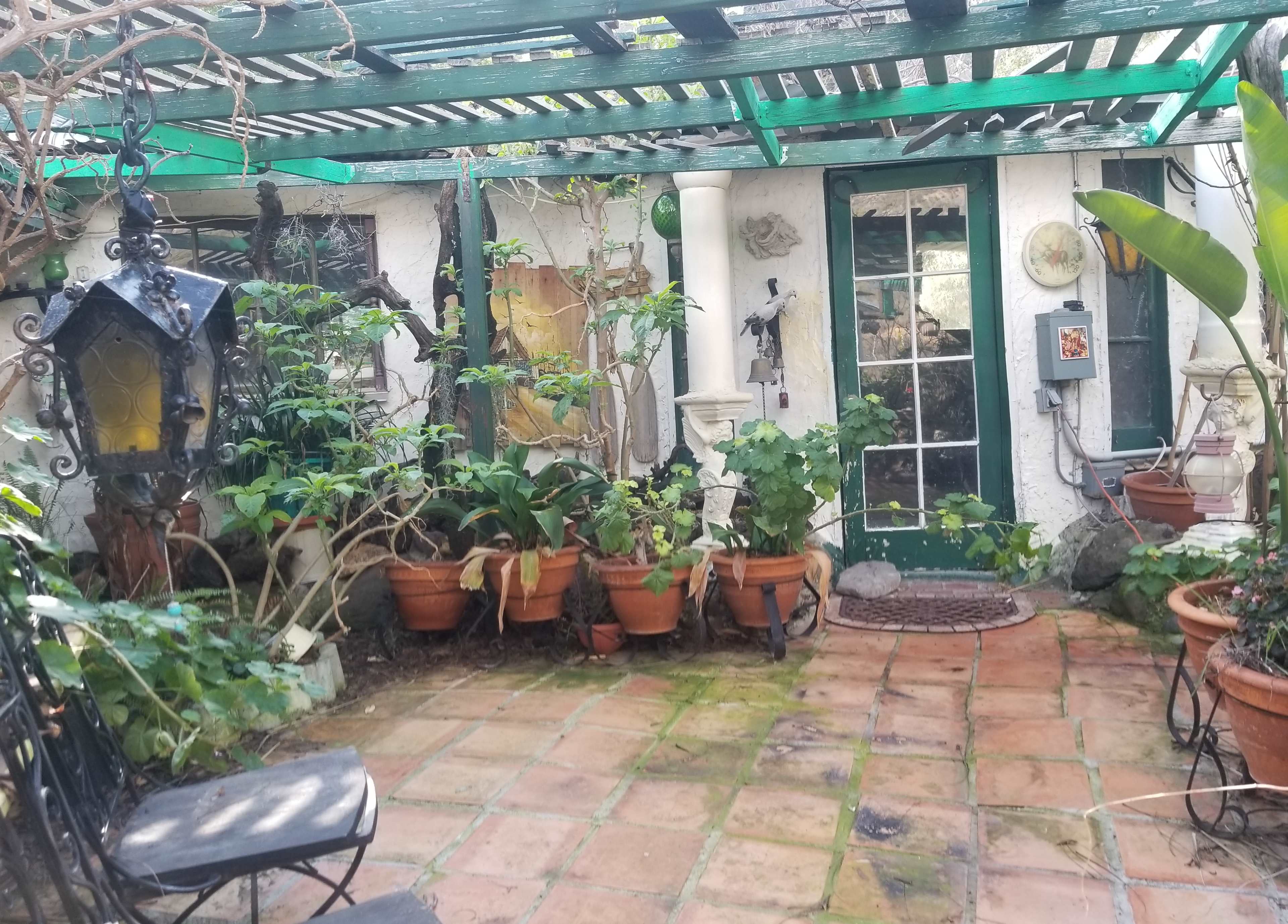 A small, sheltered courtyard with terracotta tiles, potted plants, a wrought-iron chair, and a door leading to a garden space.