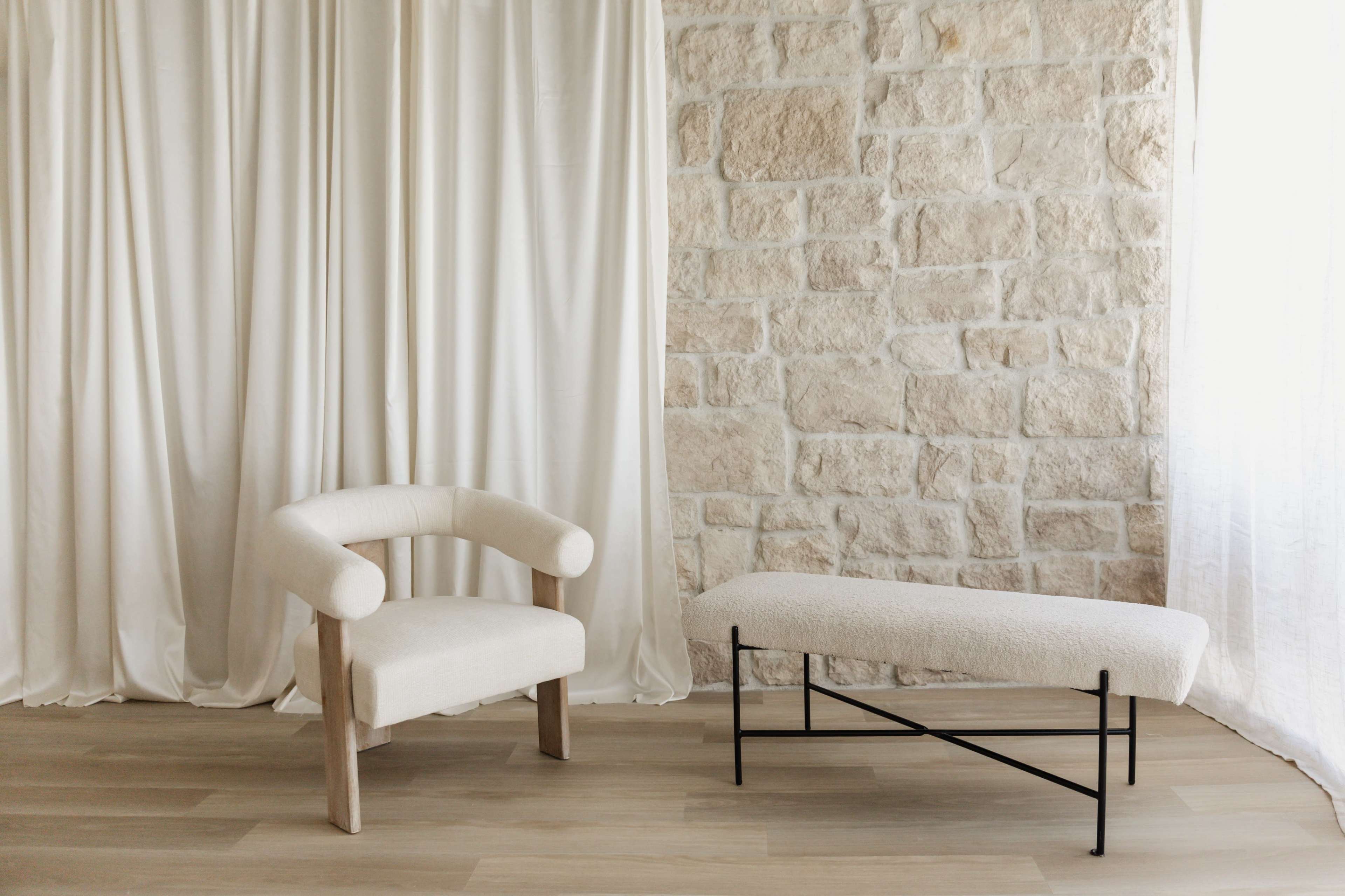 The image features a minimalist seating arrangement consisting of a curved armchair and a long bench, set against a stone wall and soft drapery.
