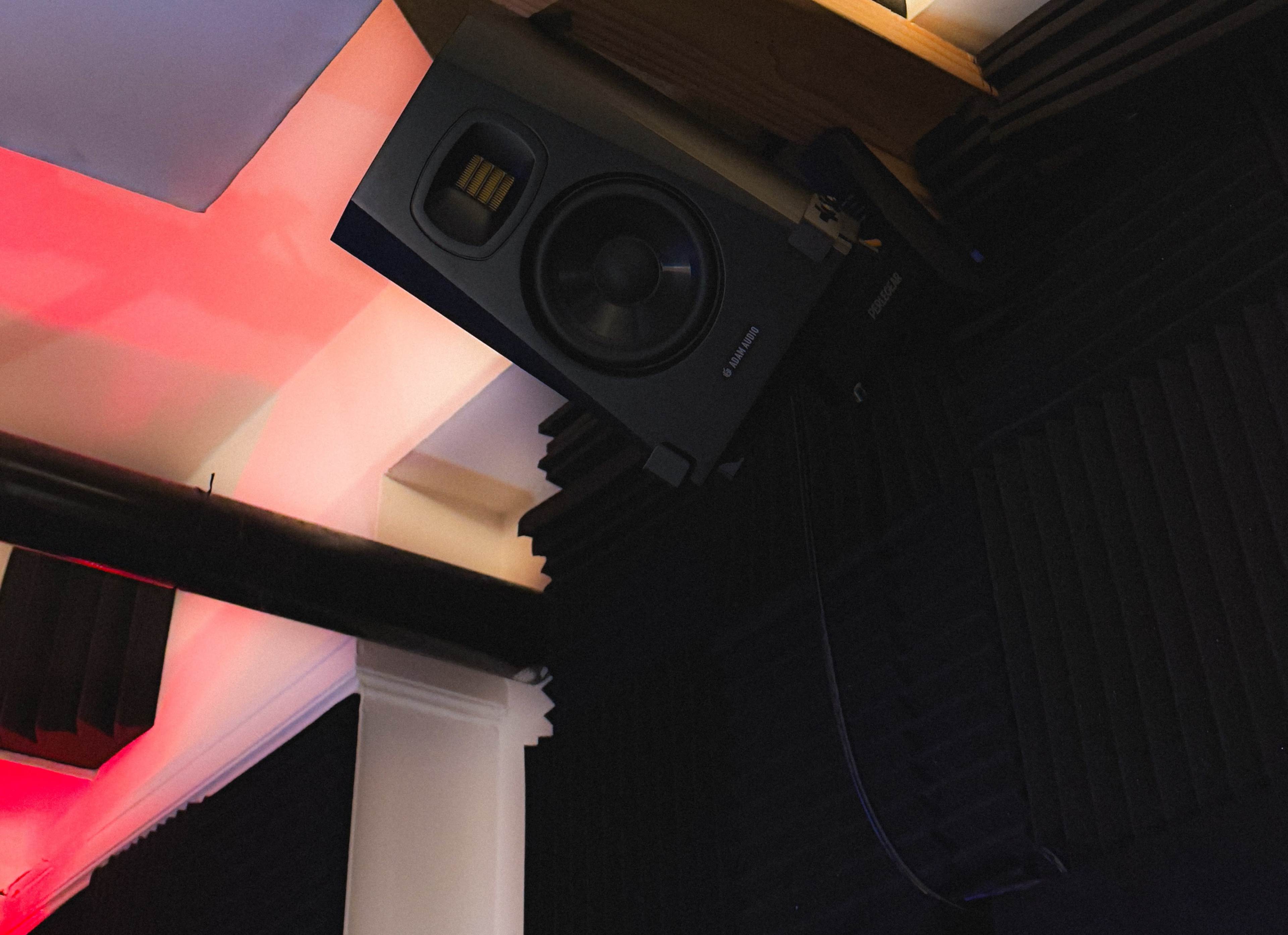 The image shows a studio monitor speaker mounted on a wall, surrounded by acoustic foam panels and colored lighting.