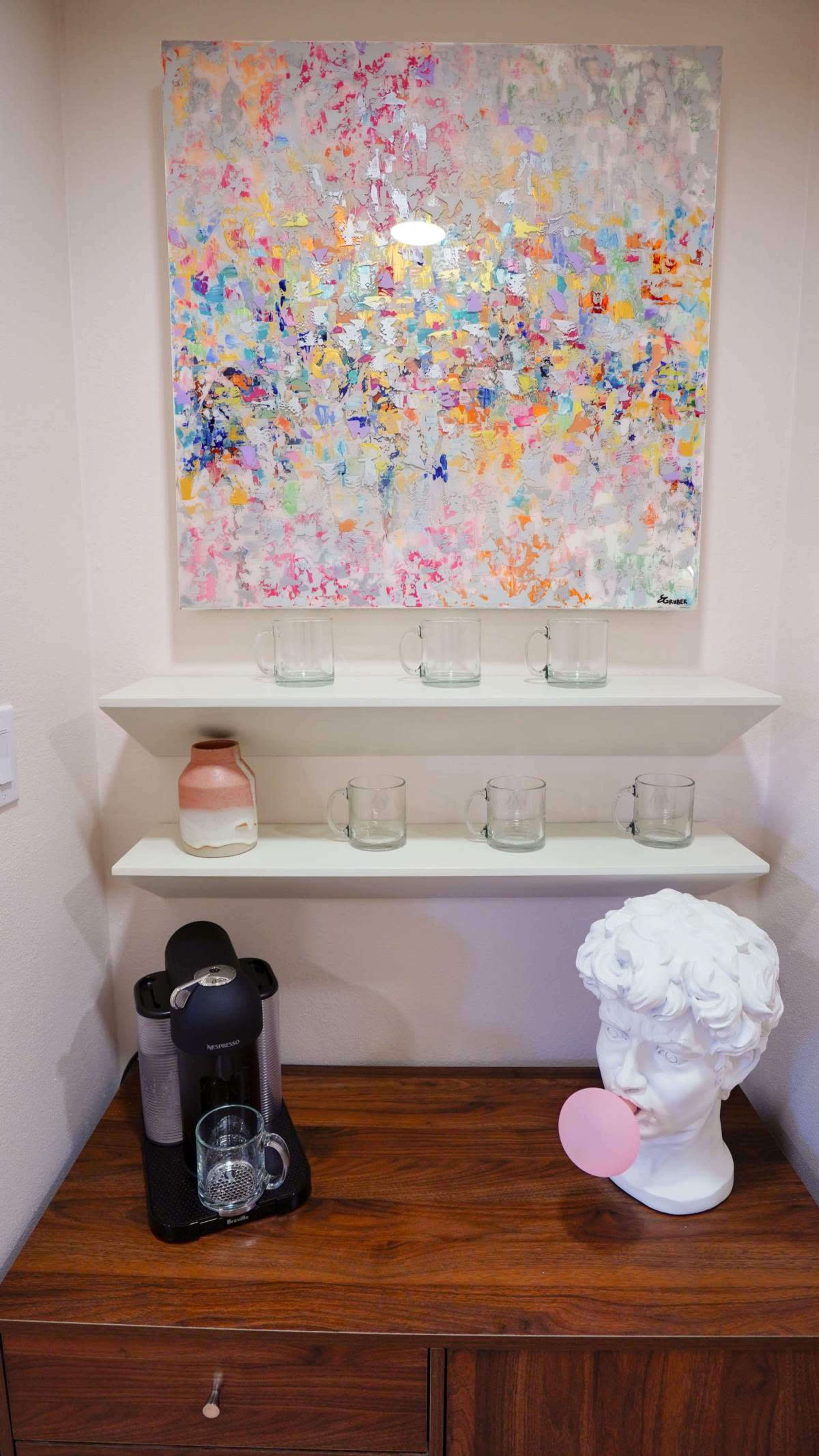 The image shows a wall-mounted shelf with several glass cups arranged on it, a pink and beige decorative vase, a coffee machine with a glass cup beside it, and a bust sculpture with a pink circle in its mouth, all beneath a colorful abstract painting.