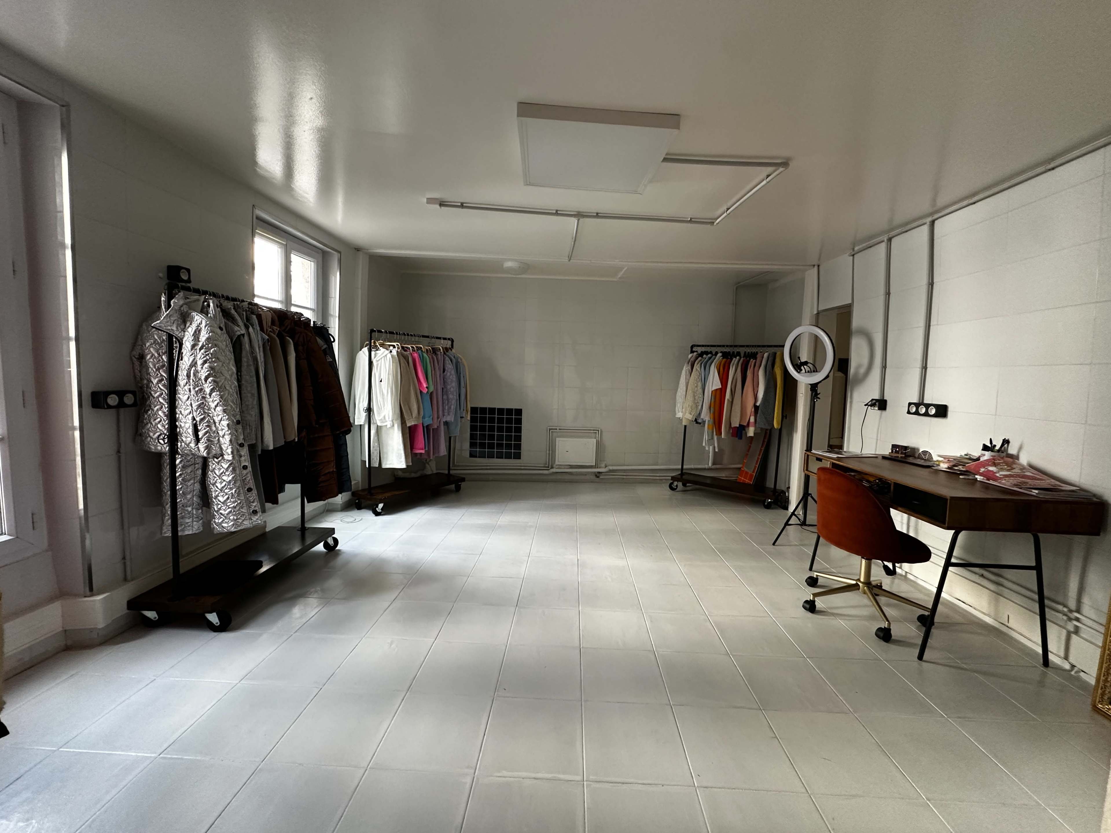 The image shows a spacious room with gray tiled flooring, featuring clothing racks on either side filled with various garments and a desk with a mirror in the corner.