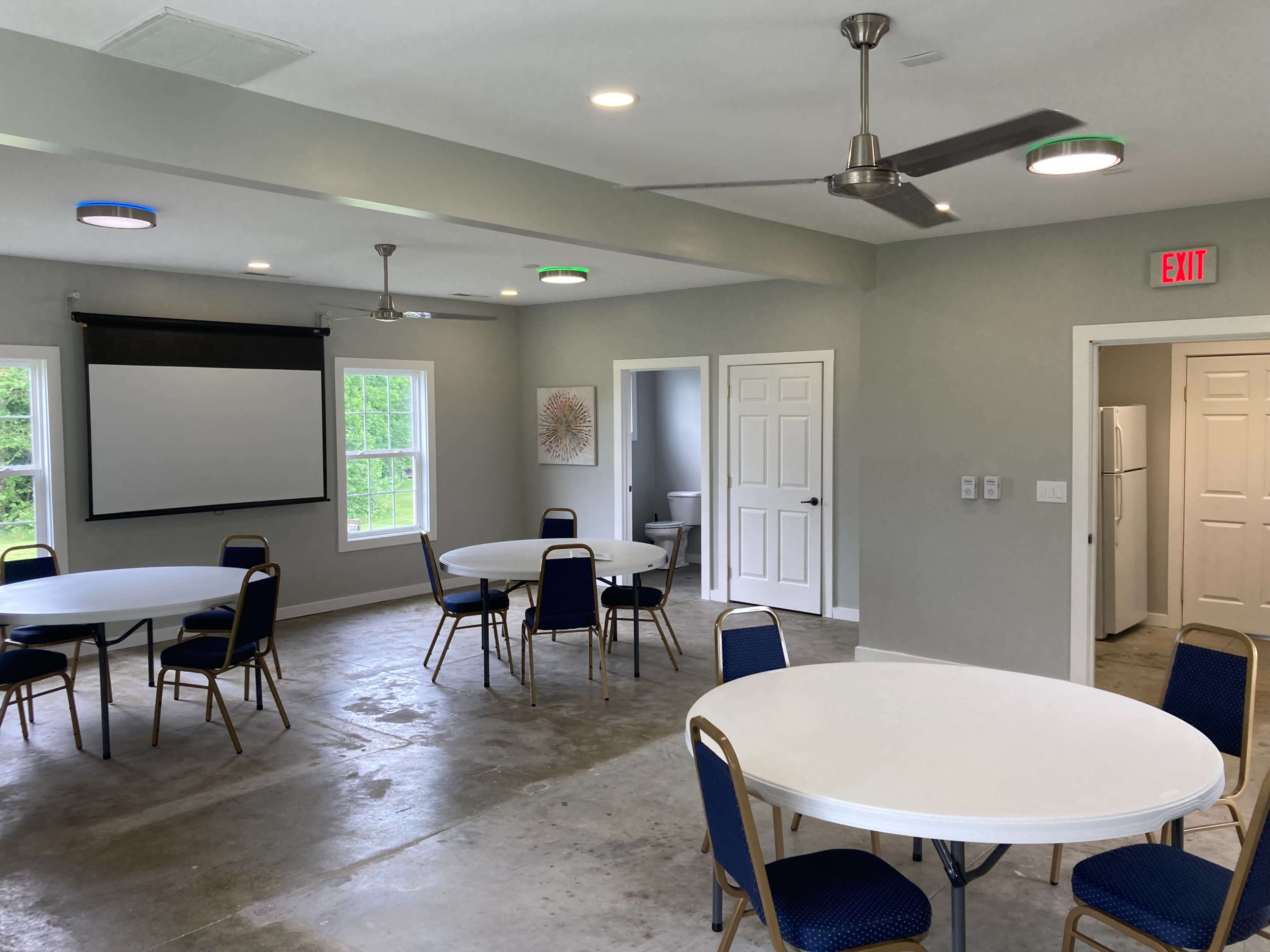 A spacious room features several round tables with blue chairs, a projector screen, and a kitchenette area in the background.