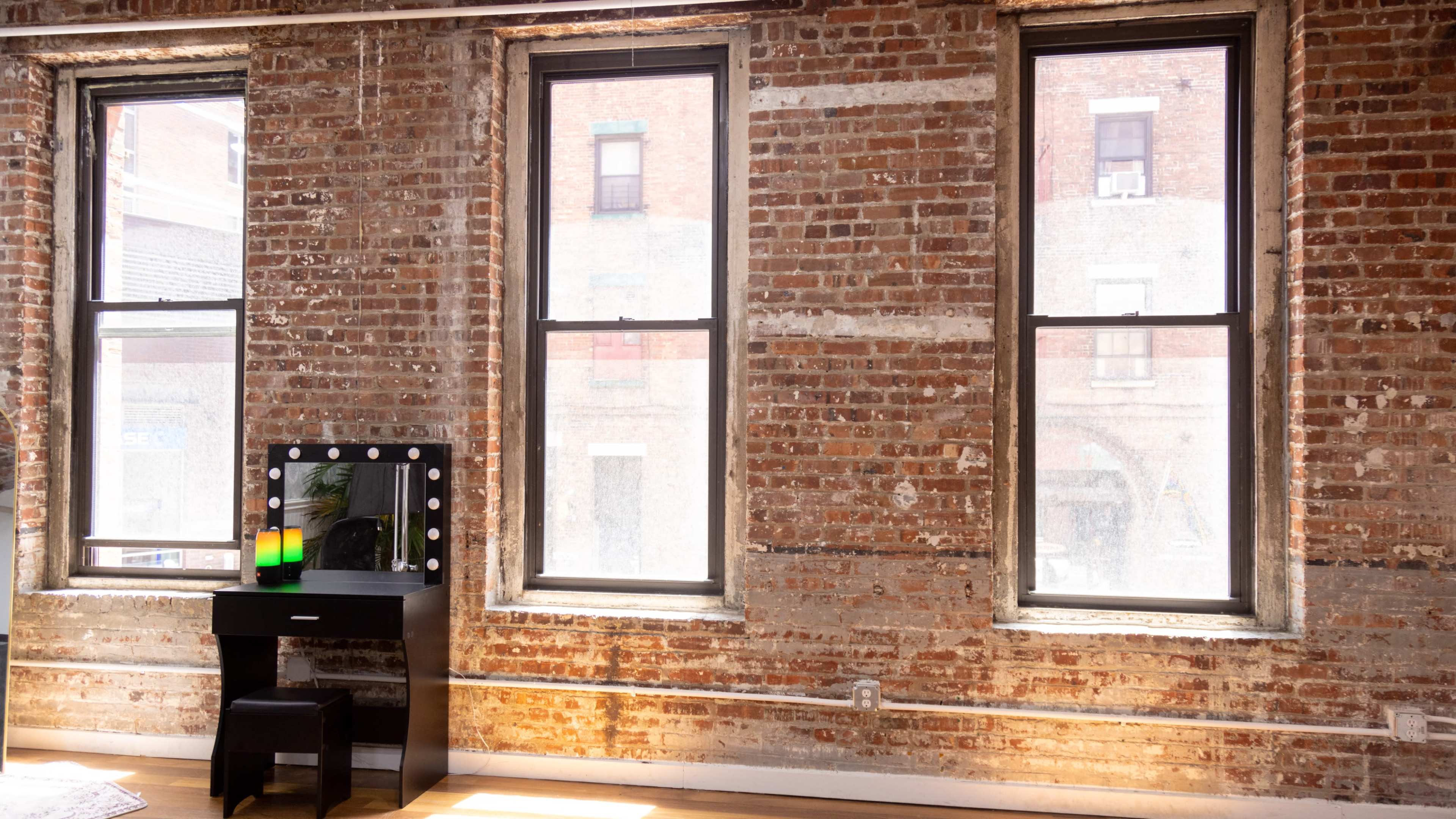 A room with three large windows framed by exposed brick walls and a black vanity mirror with lights in front of it.