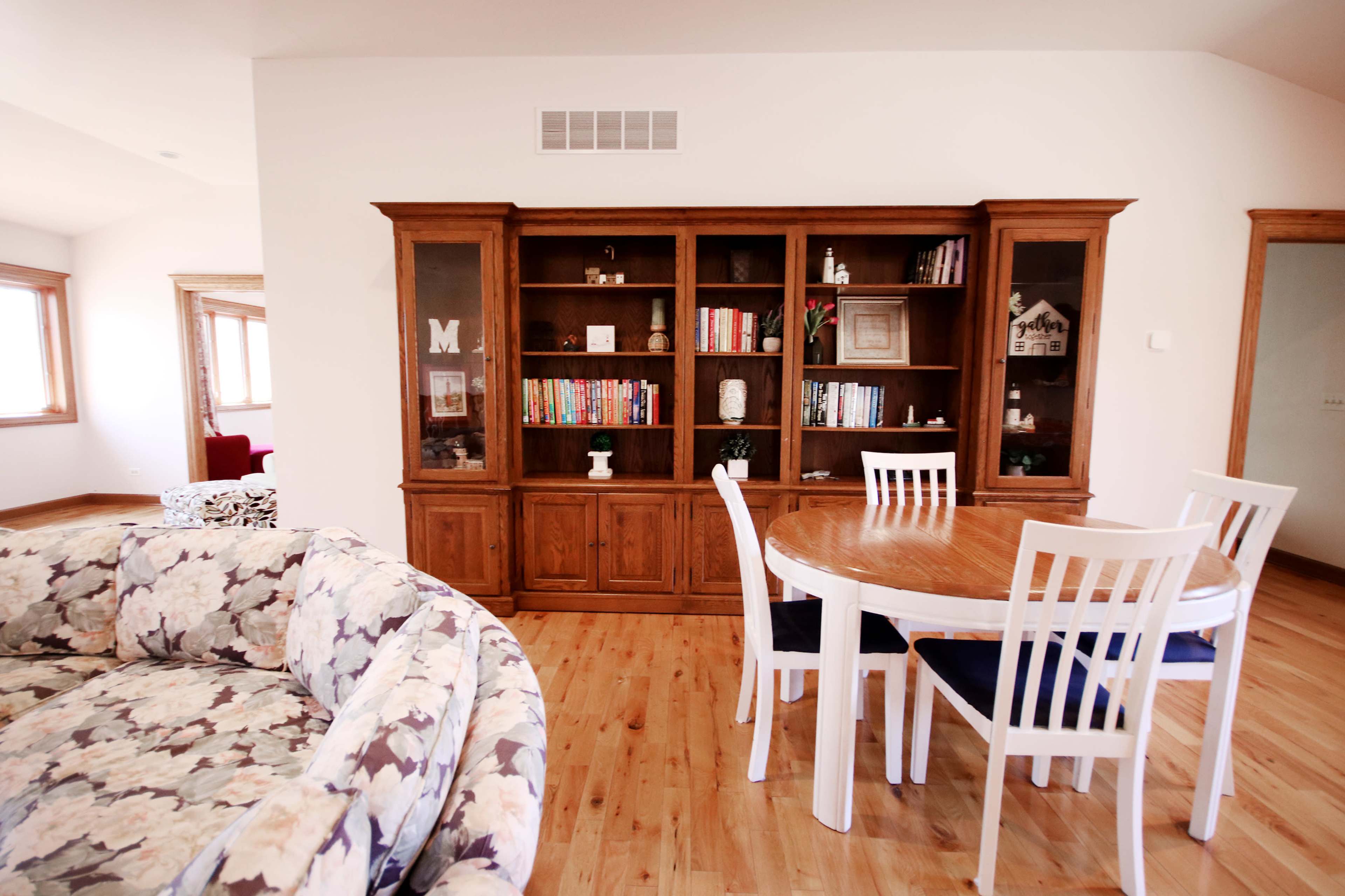A well-lit room with a wooden bookshelf filled with books and a round dining table surrounded by white chairs.