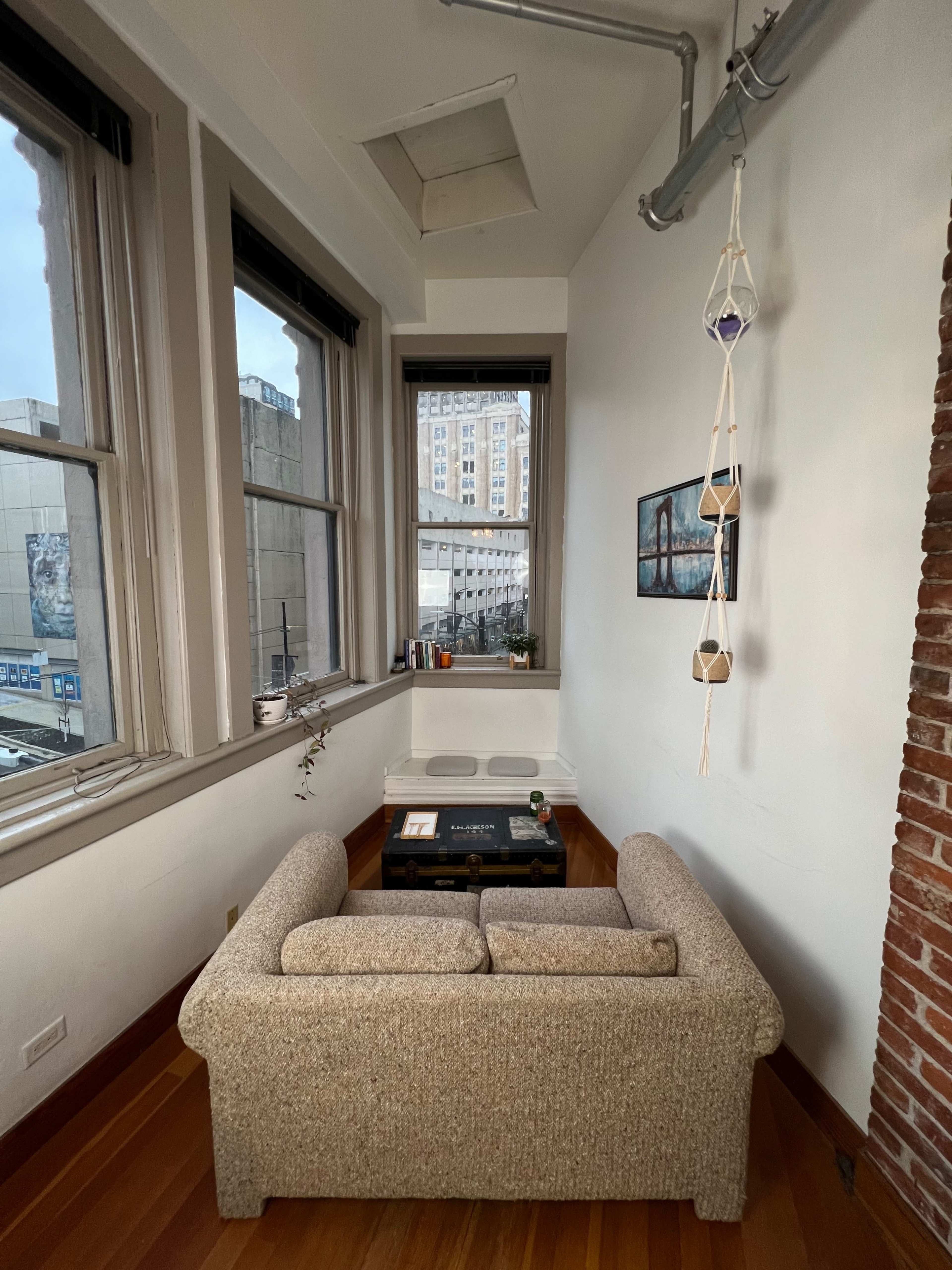 A cozy corner of an apartment features a beige sofa, a small coffee table, and large windows with a view of a cityscape.