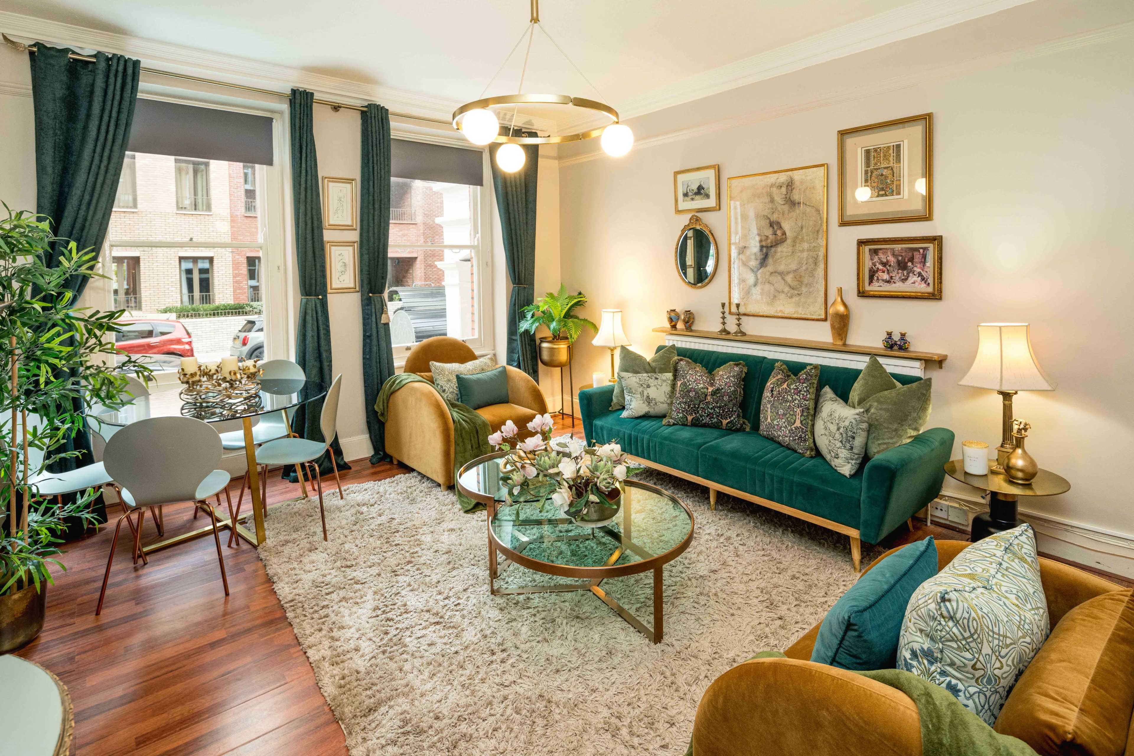 The image shows a well-decorated living room with a green velvet sofa, a round glass coffee table, and a combination of armchairs and dining chairs arranged near a window.