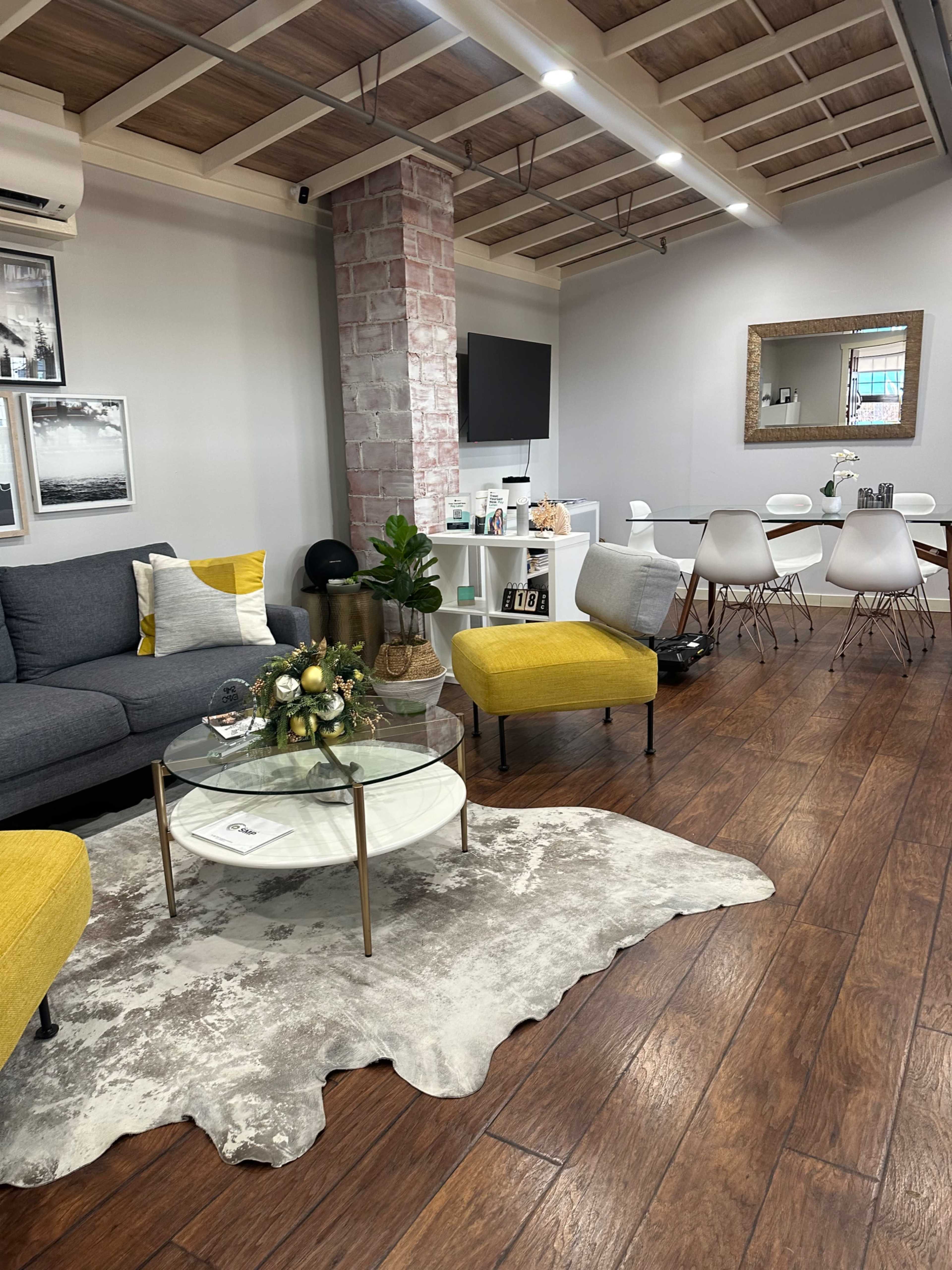 The image shows a modern living area featuring a gray sofa with yellow accent chairs, a coffee table on a patterned rug, a TV mounted on the wall, and a dining area with white chairs and a mirror.