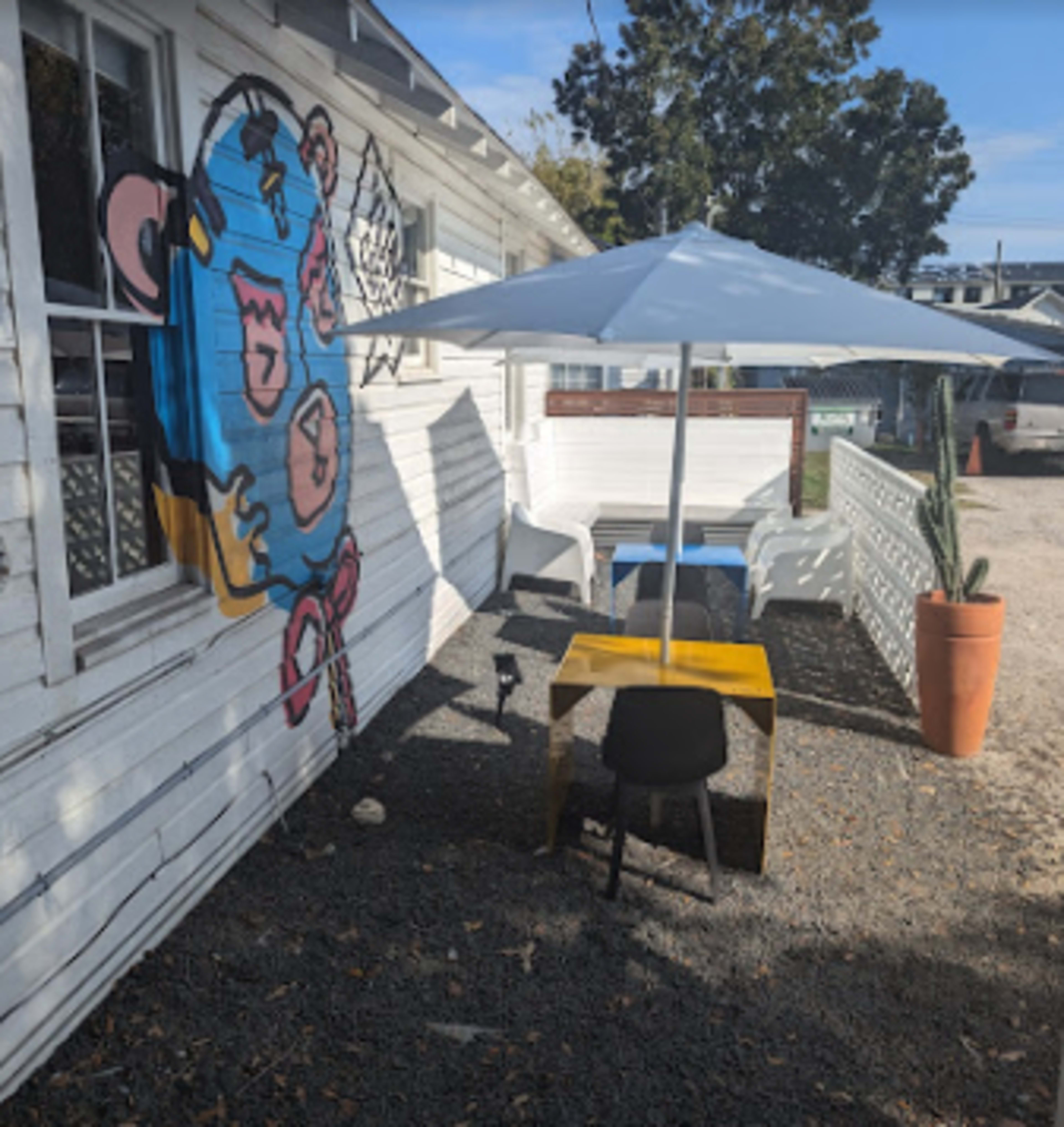 A small outdoor seating area with a yellow table, black chairs, and a white umbrella next to a building featuring a colorful mural.