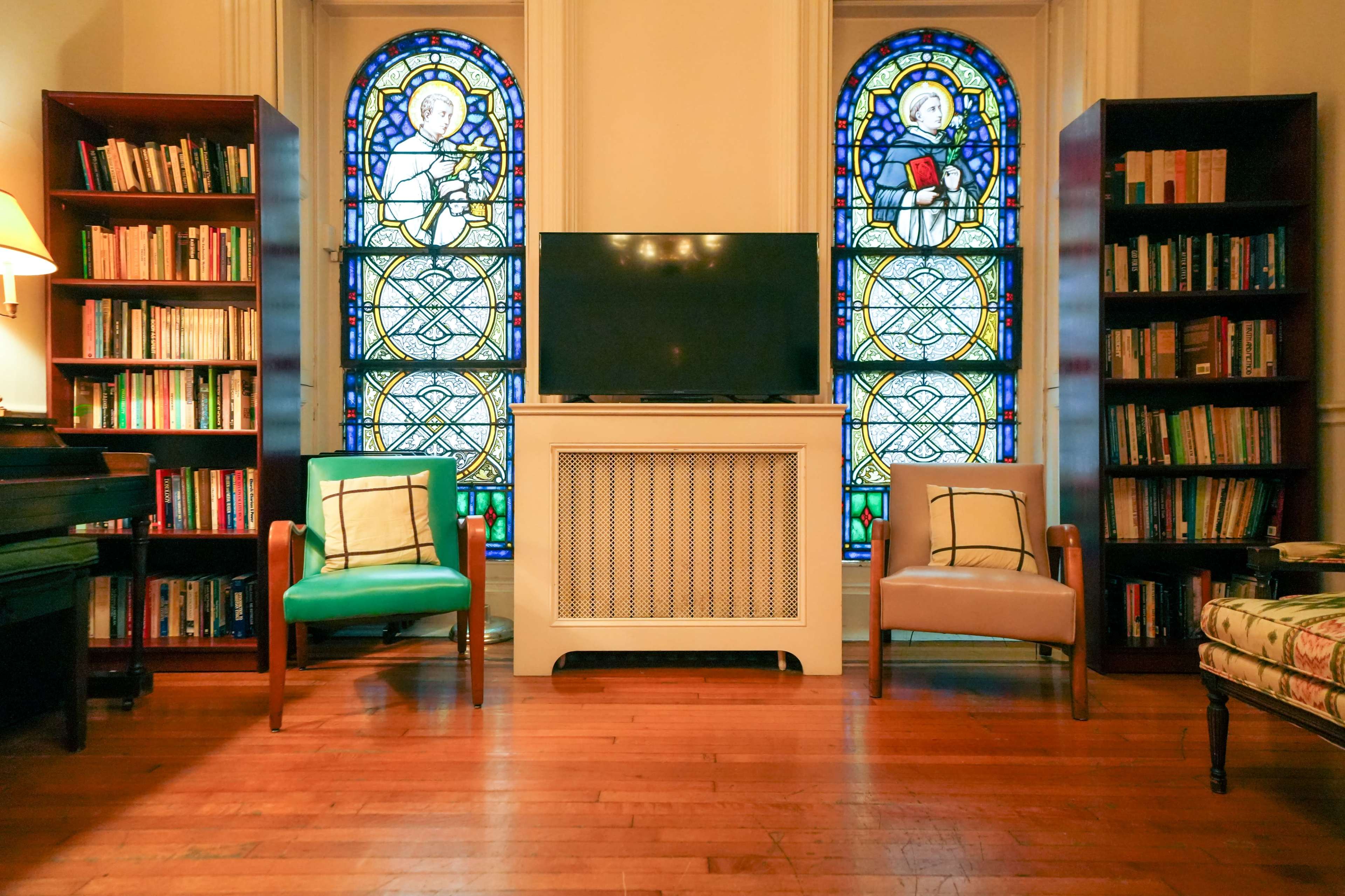 The room features two chairs, a television on a white cabinet, and bookshelves filled with books, set against the backdrop of stained glass windows.