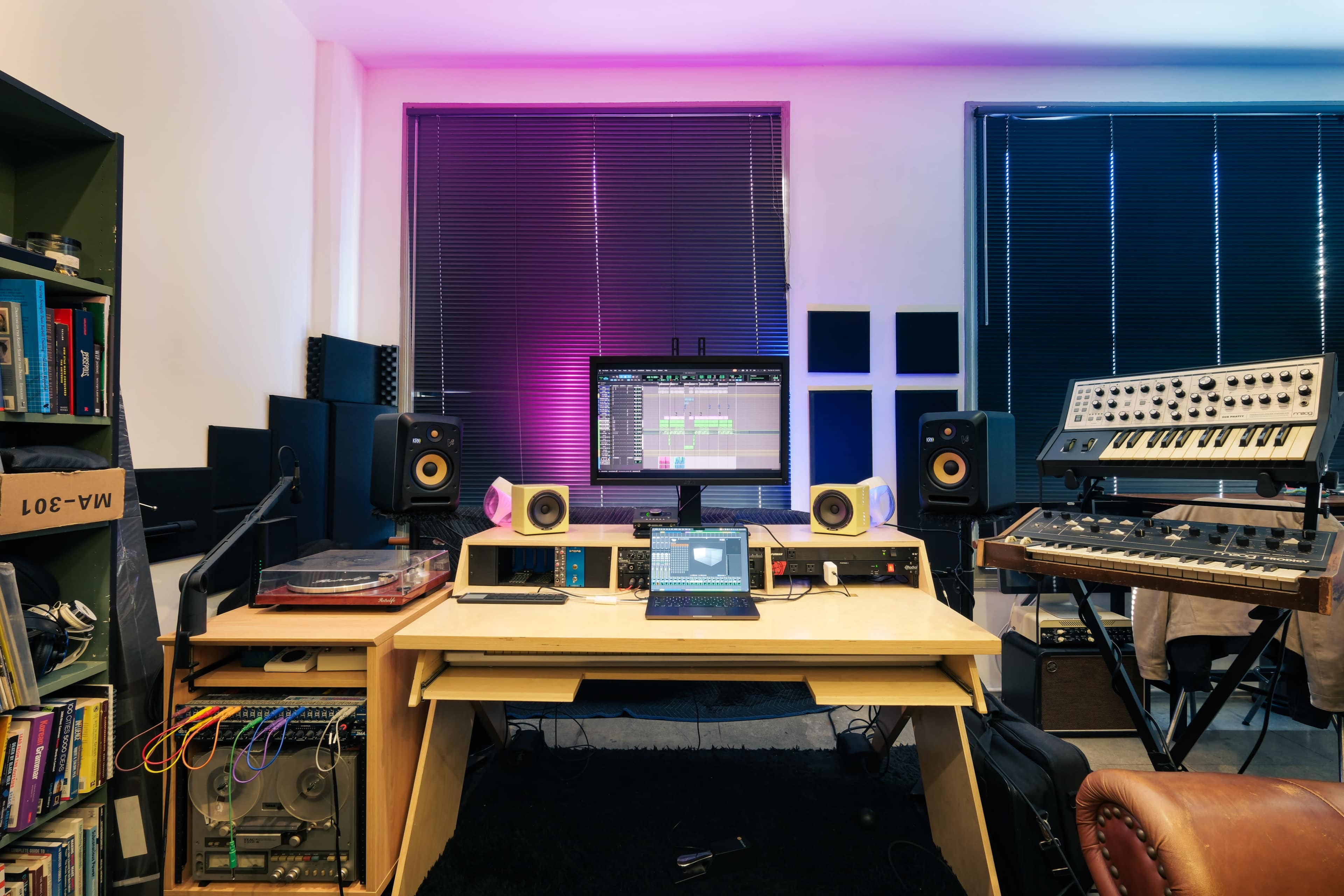 The image shows a music production setup featuring a computer monitor displaying software, several audio speakers, a synthesizer, and a desk with various equipment in a well-lit room.