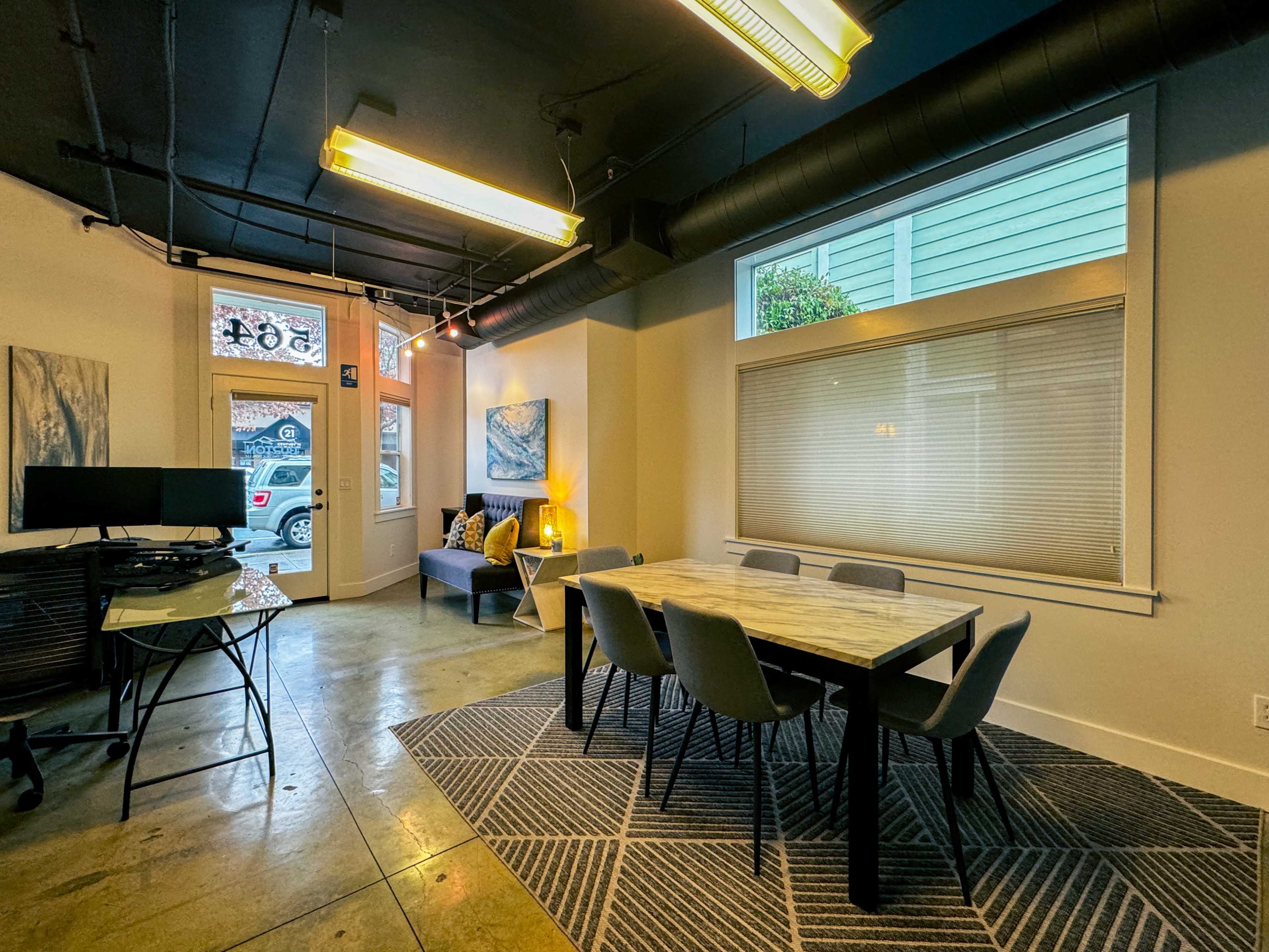 A modern workspace with a large table, several chairs, a couch, and large windows letting in natural light.