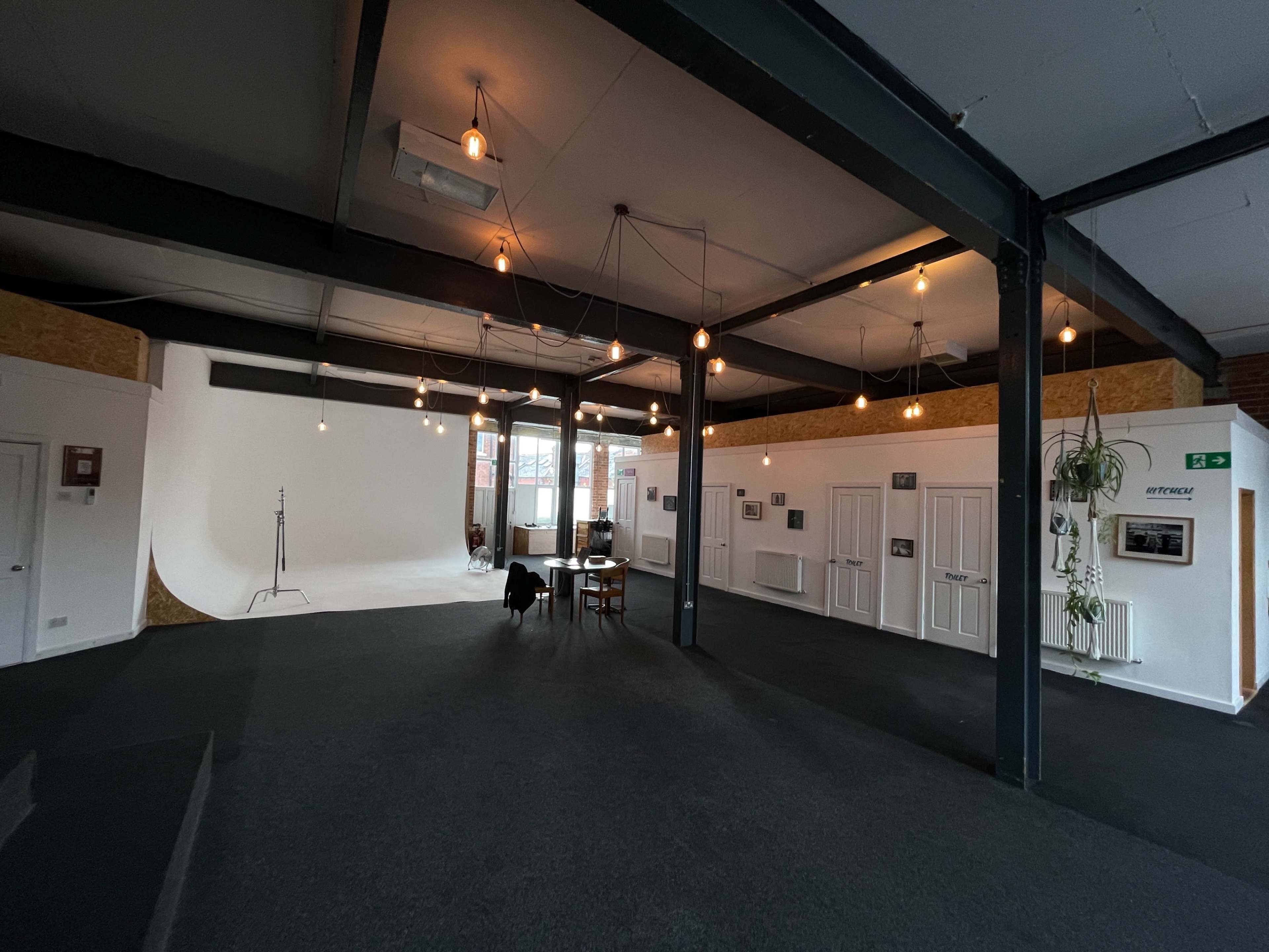 A spacious studio with wooden paneling, a white backdrop, and several hanging light bulbs, accompanied by a table and chairs against the walls.