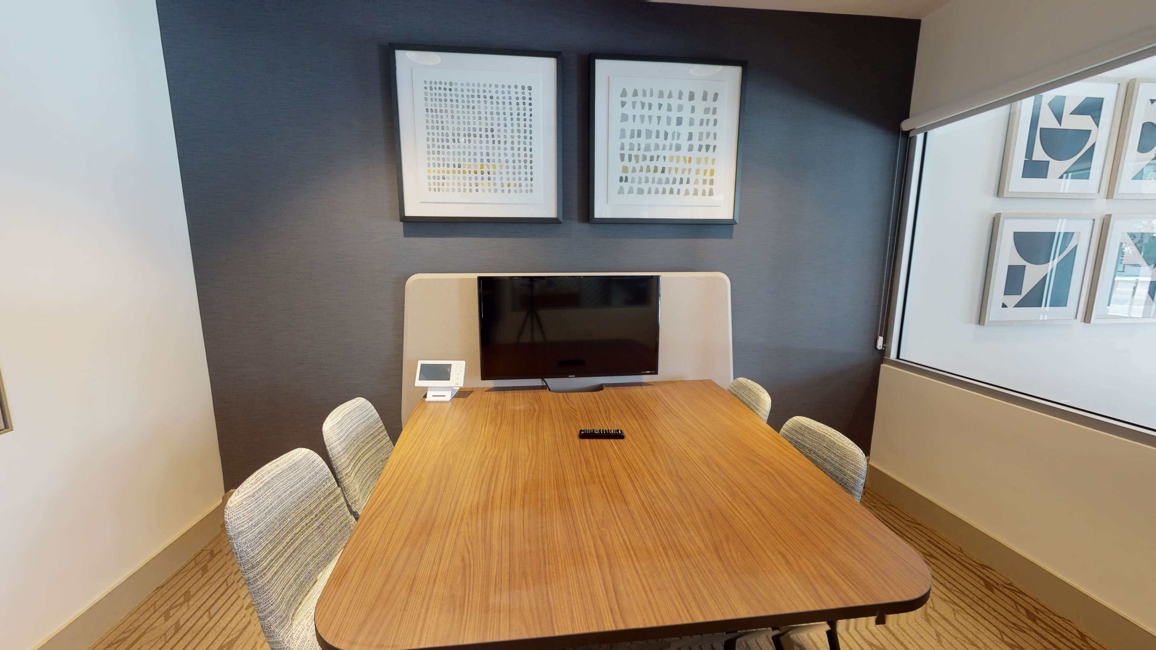 A conference room with a wooden table surrounded by four chairs, featuring a wall-mounted television and framed artwork above.