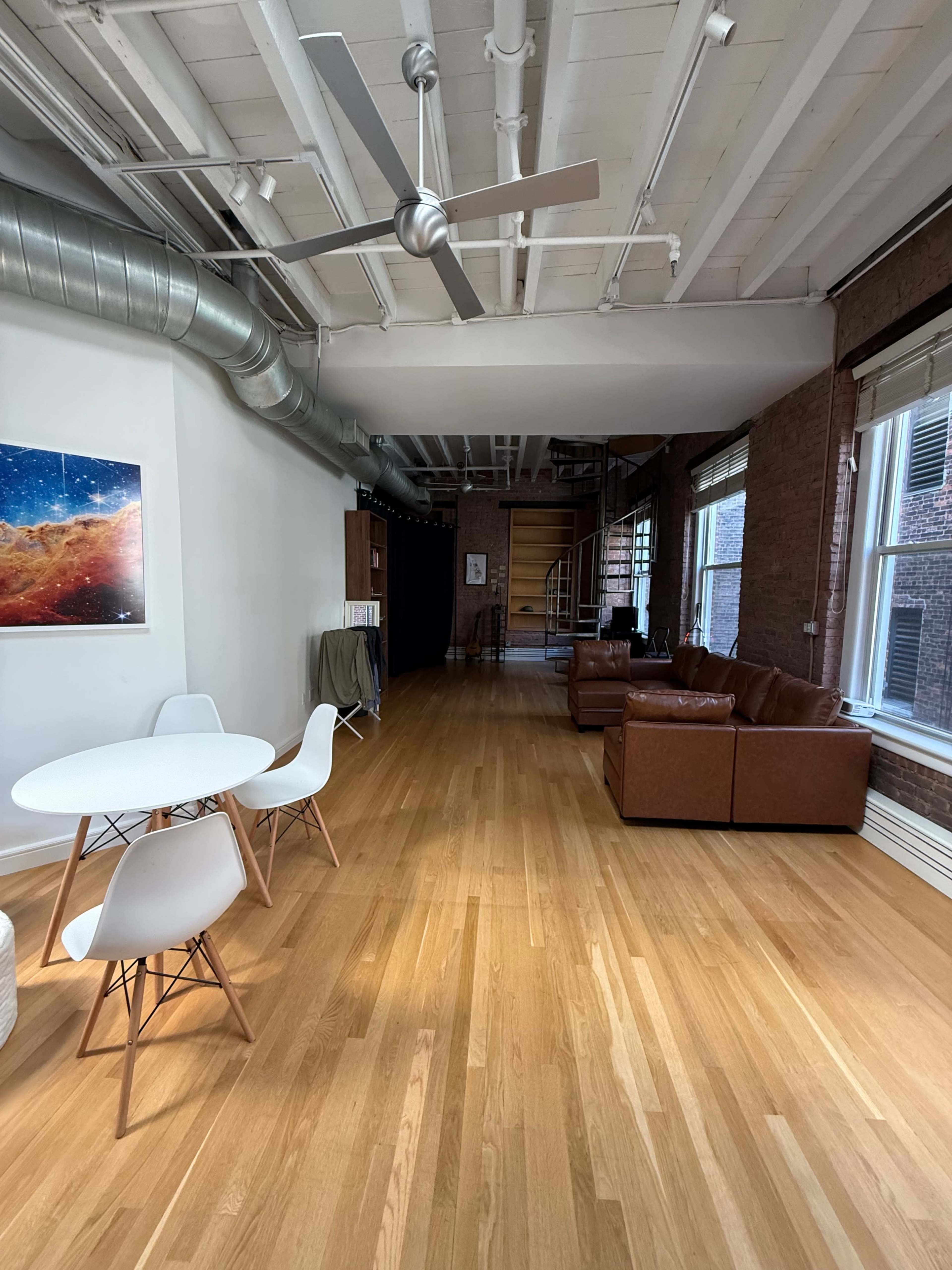 The image shows a spacious, modern interior with wooden flooring, a circular dining table with white chairs, brown leather couches, and a spiral staircase in the background.