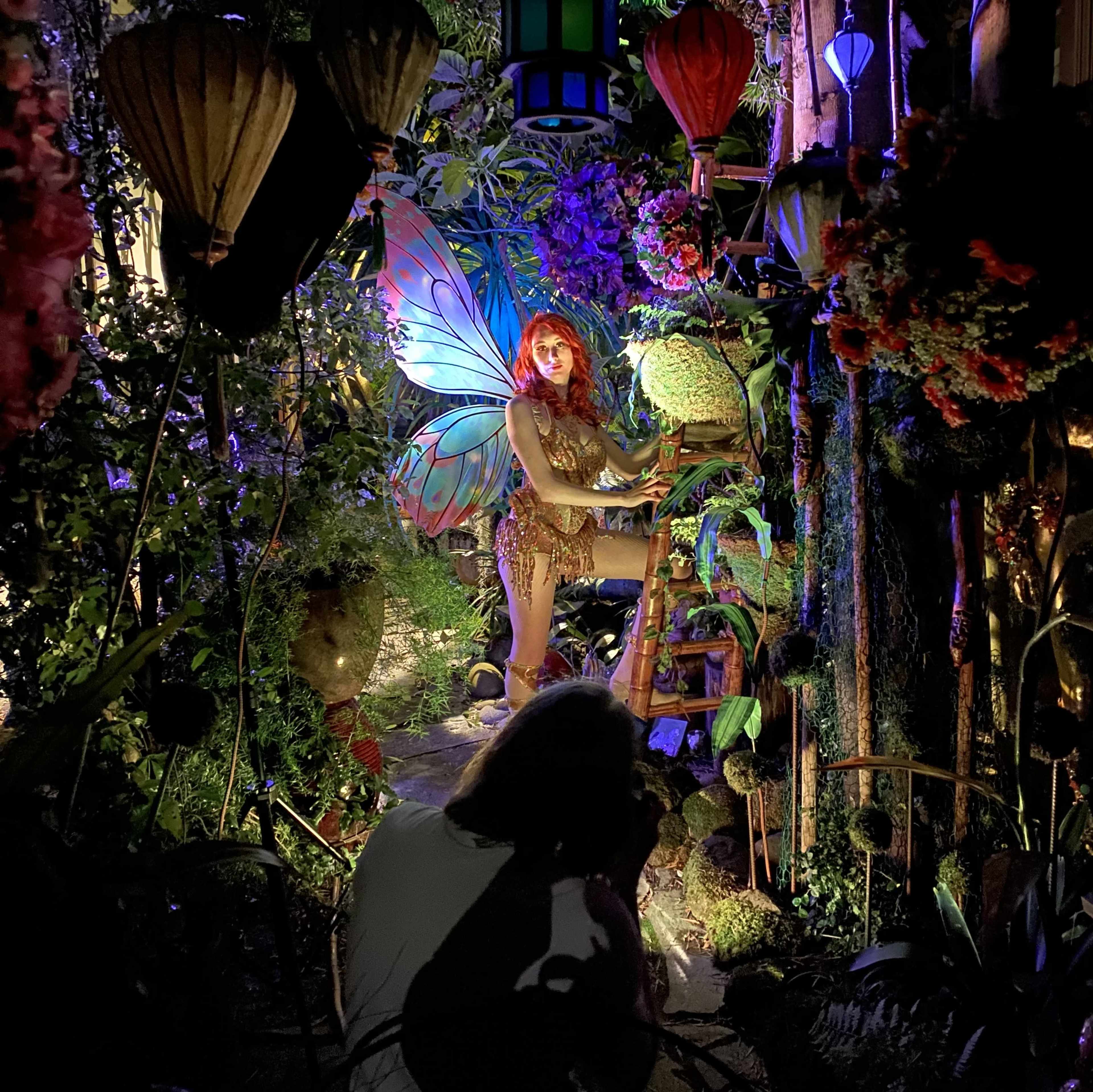 A person takes a photograph of a model with butterfly wings posed amidst vibrant decorations in a garden setting.