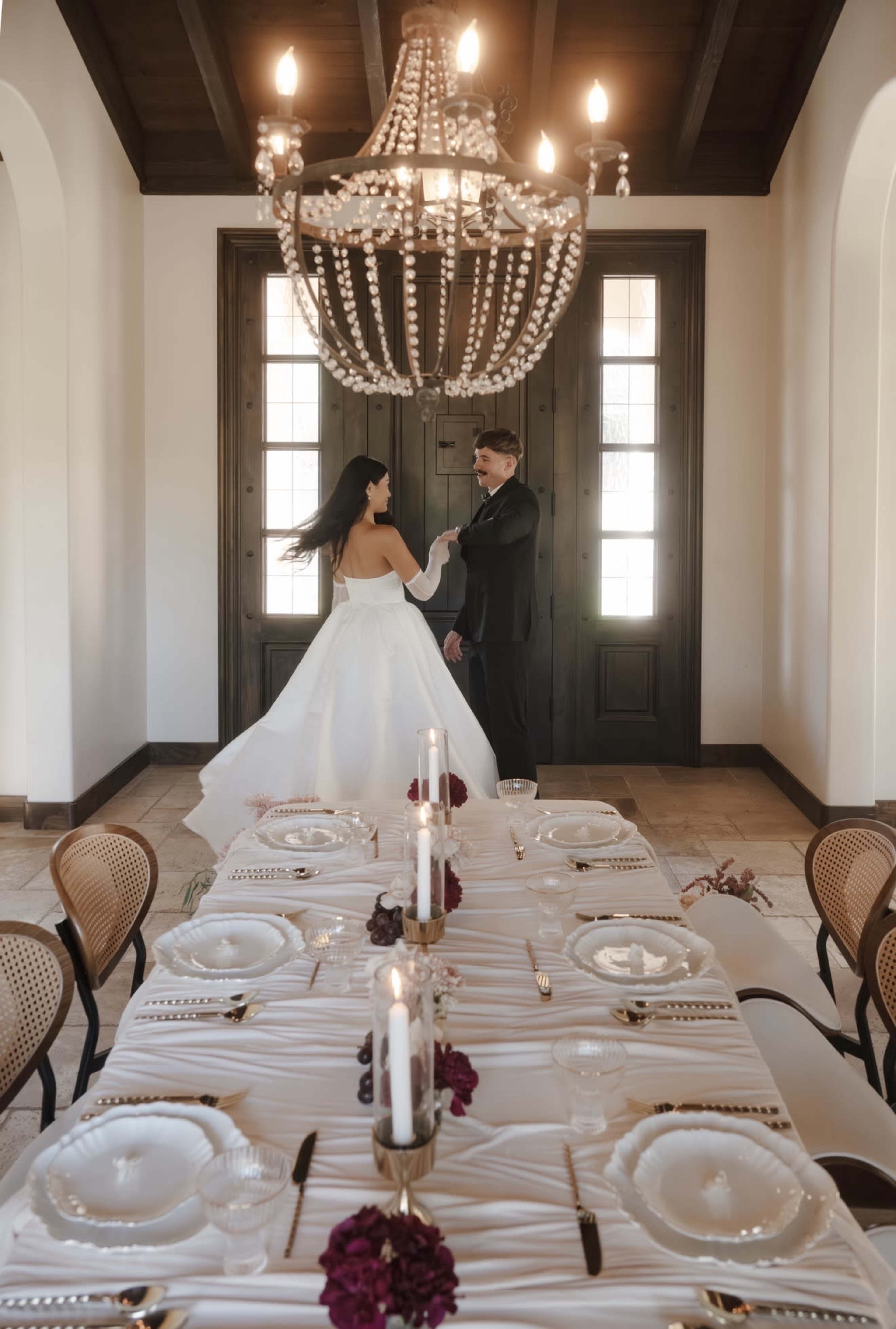 A couple dressed in formal attire stands in a beautifully decorated dining room with a large chandelier above and a table set for an elegant meal.
