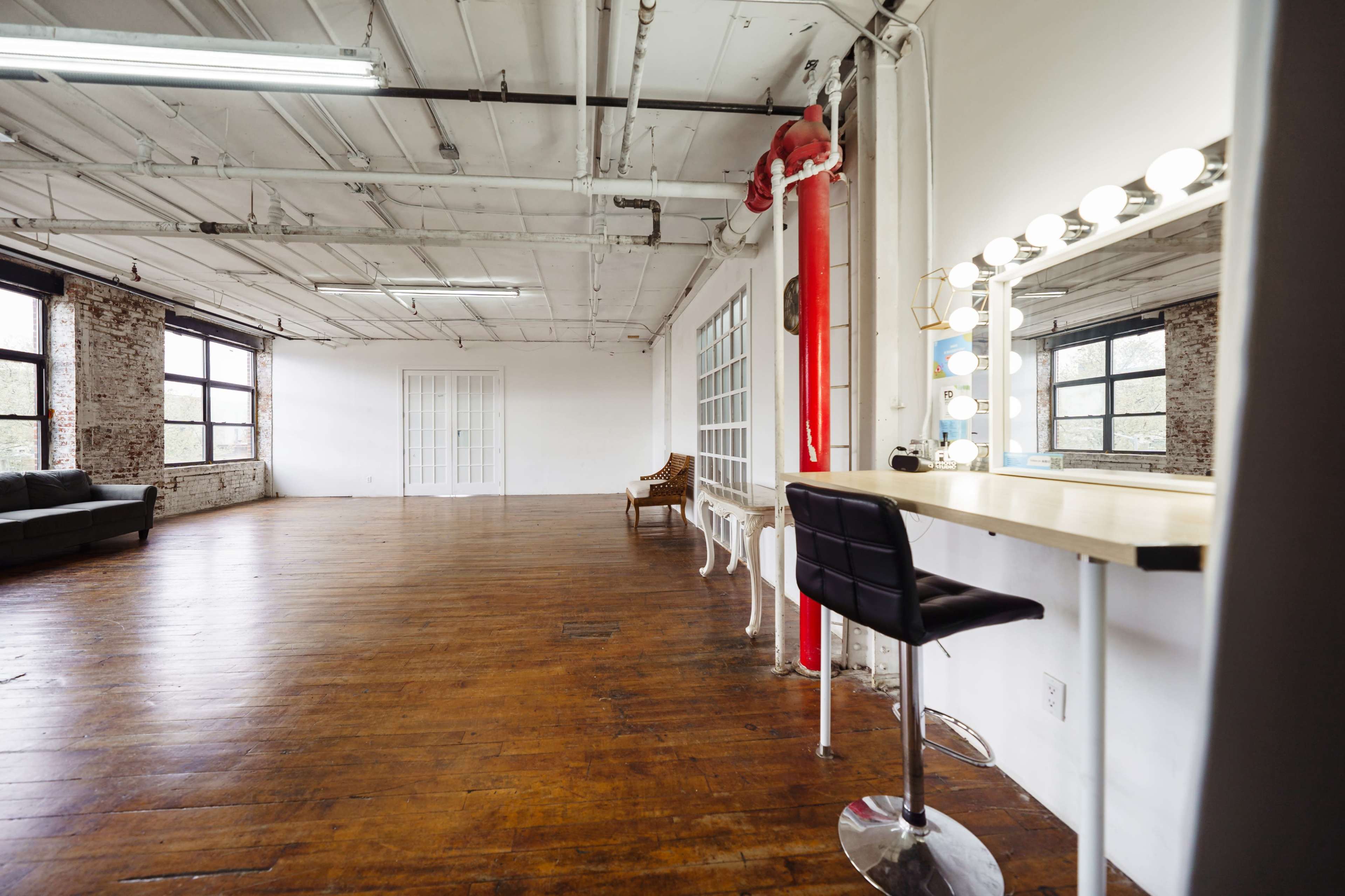 A spacious, industrial-style room with wooden flooring, large windows, and a makeup station with a spotlight mirror.