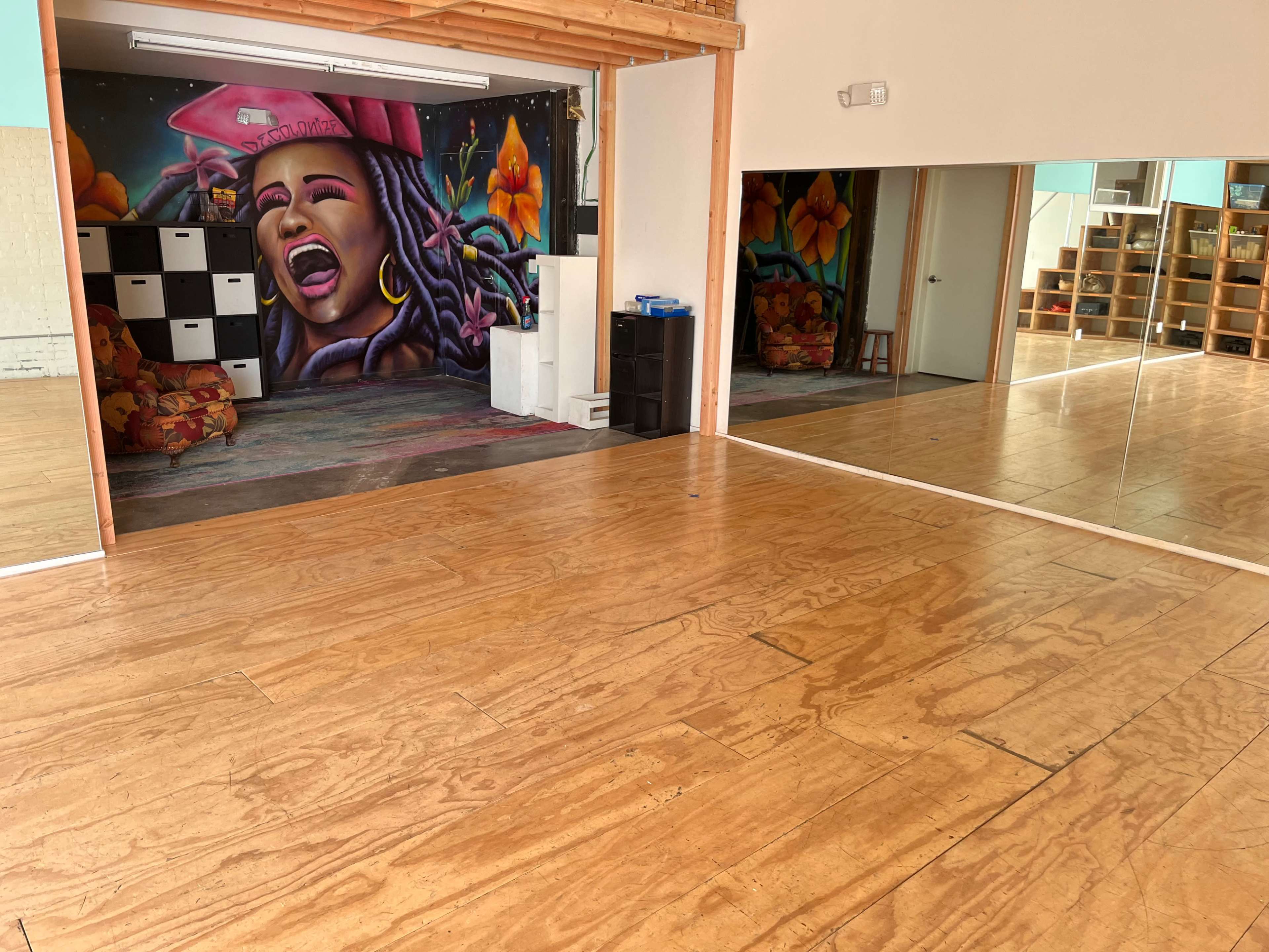 A spacious room with wooden flooring, featuring a colorful mural on one wall and mirrored surfaces reflecting the interior.