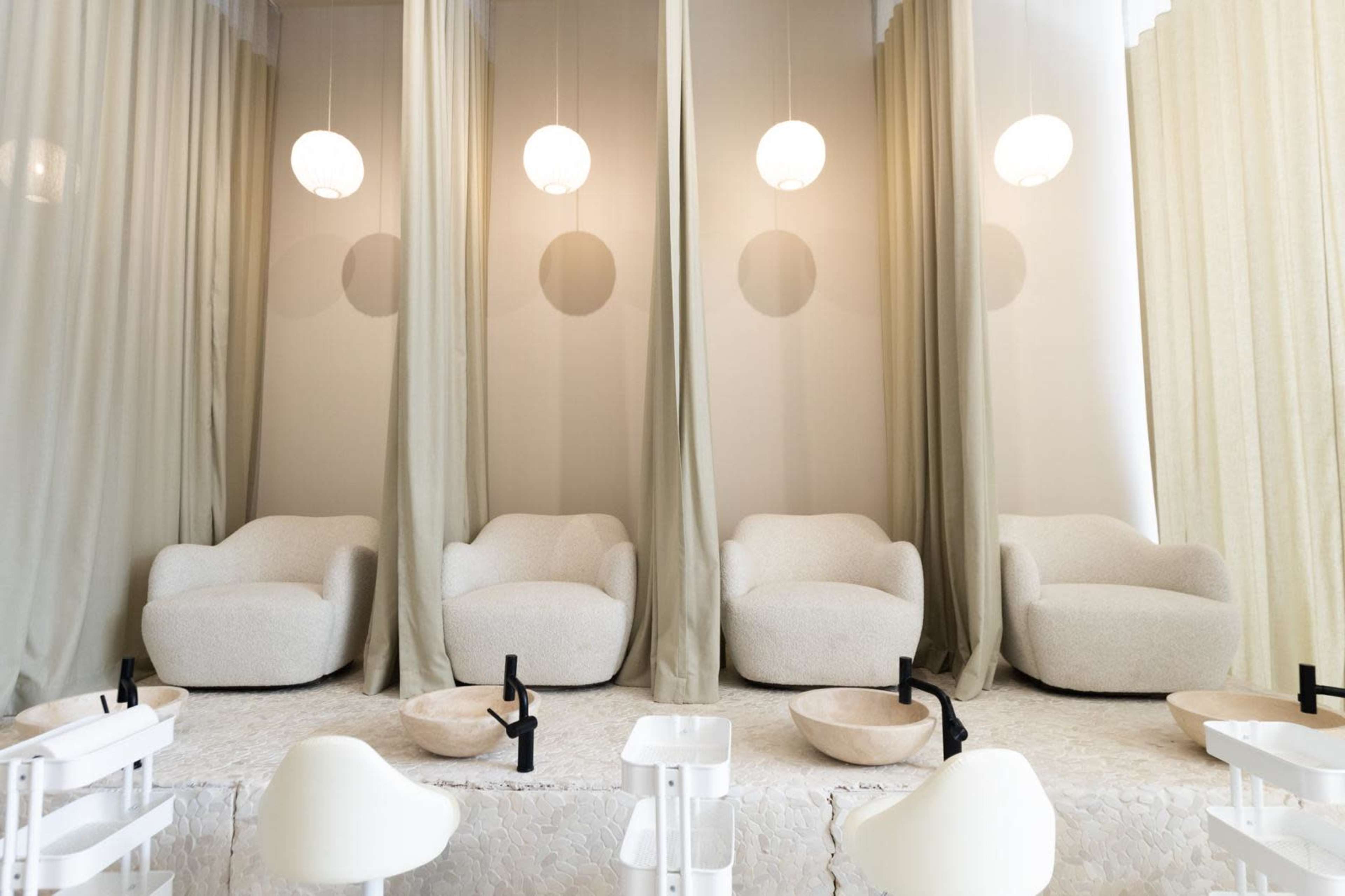 The image shows a minimalist nail salon with four cream-colored chairs separated by light curtains and pendant lighting overhead.