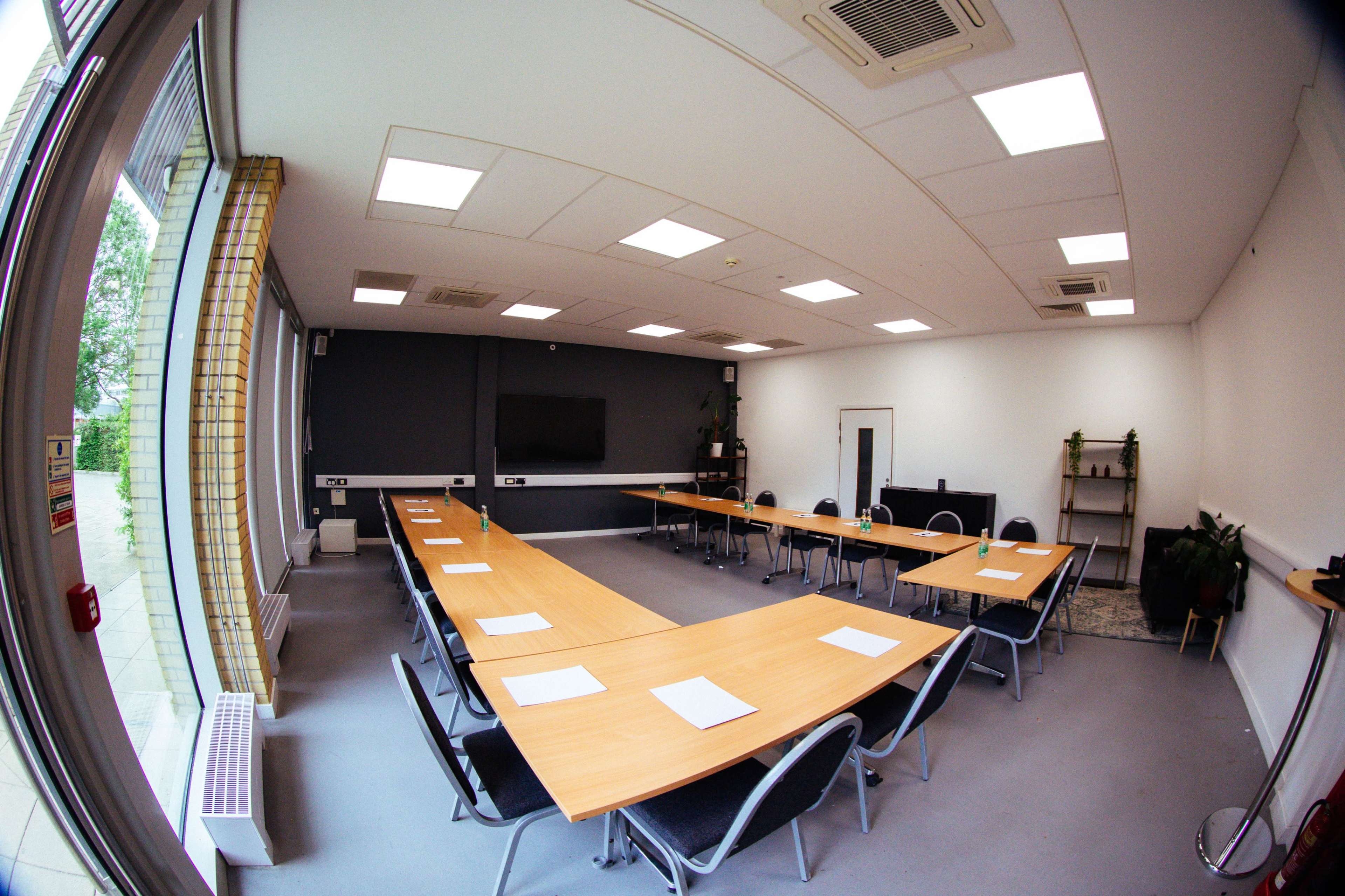 A well-lit conference room features a long arrangement of tables with chairs and a large screen at one end.