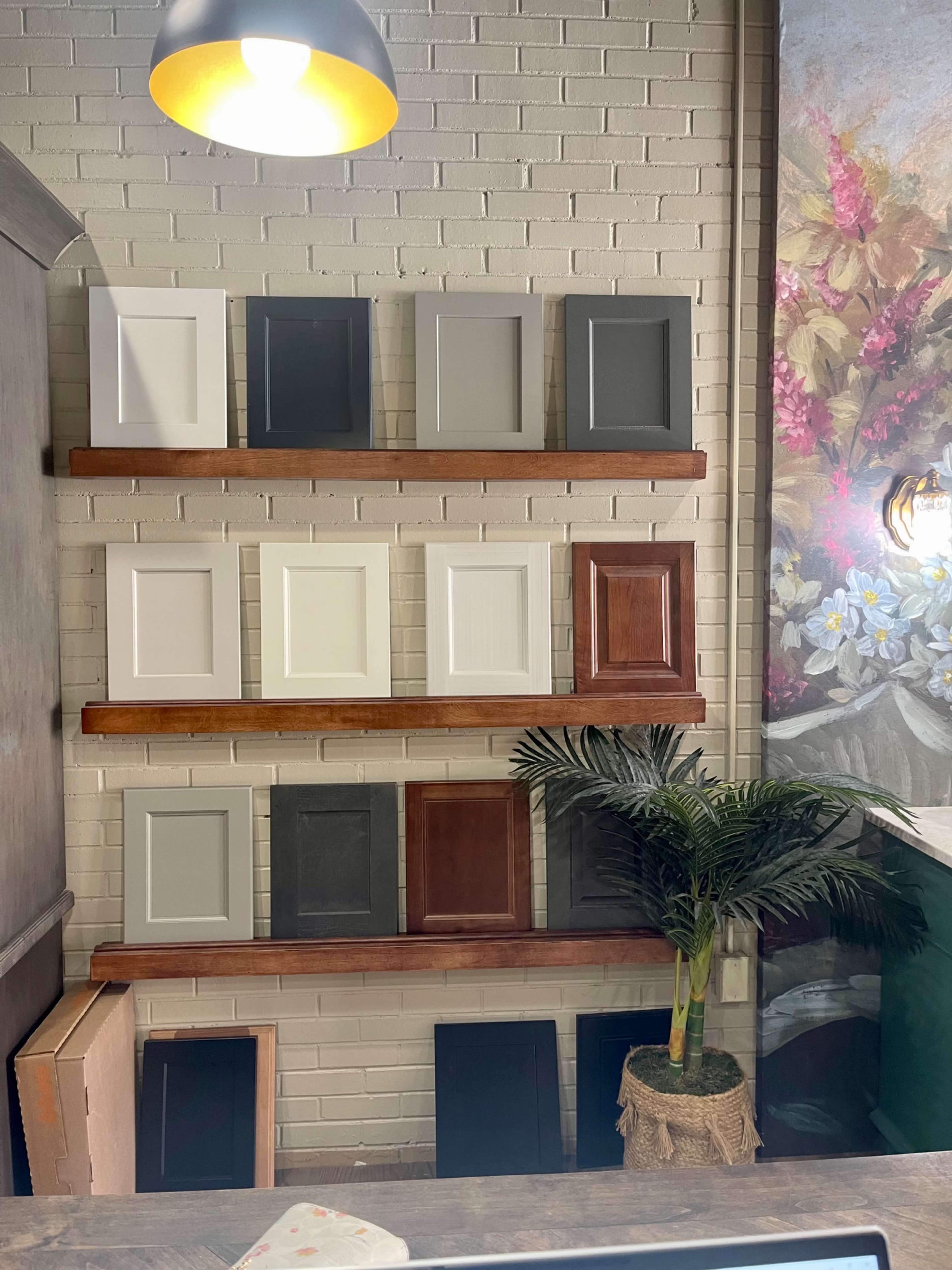 The image shows a display of various cabinet door samples arranged on wooden shelves against a white brick wall, with a potted plant in the foreground.