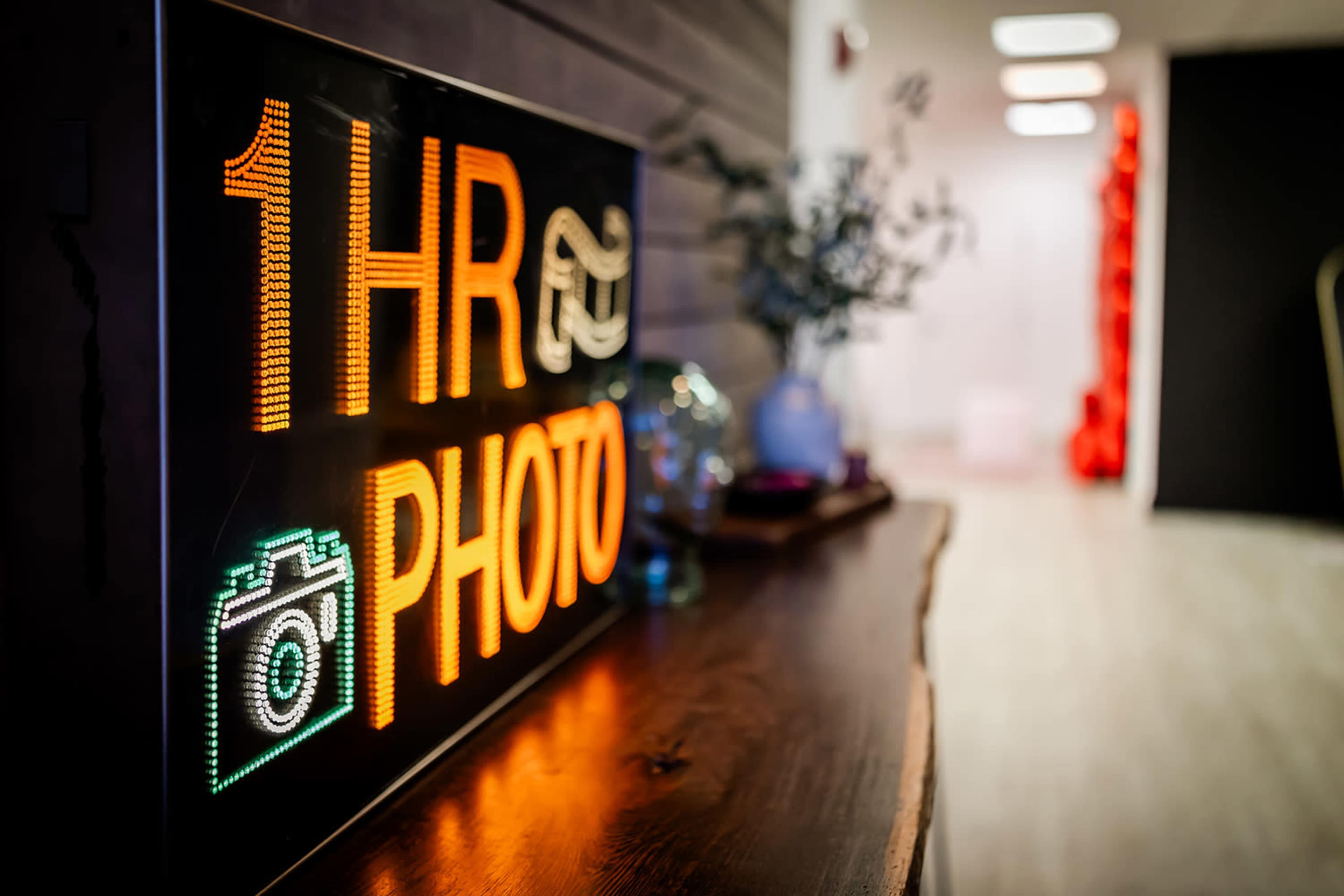The image shows a brightly lit sign displaying "1 HR PHOTO" in orange and white letters, placed on a wooden table in a modern interior space.