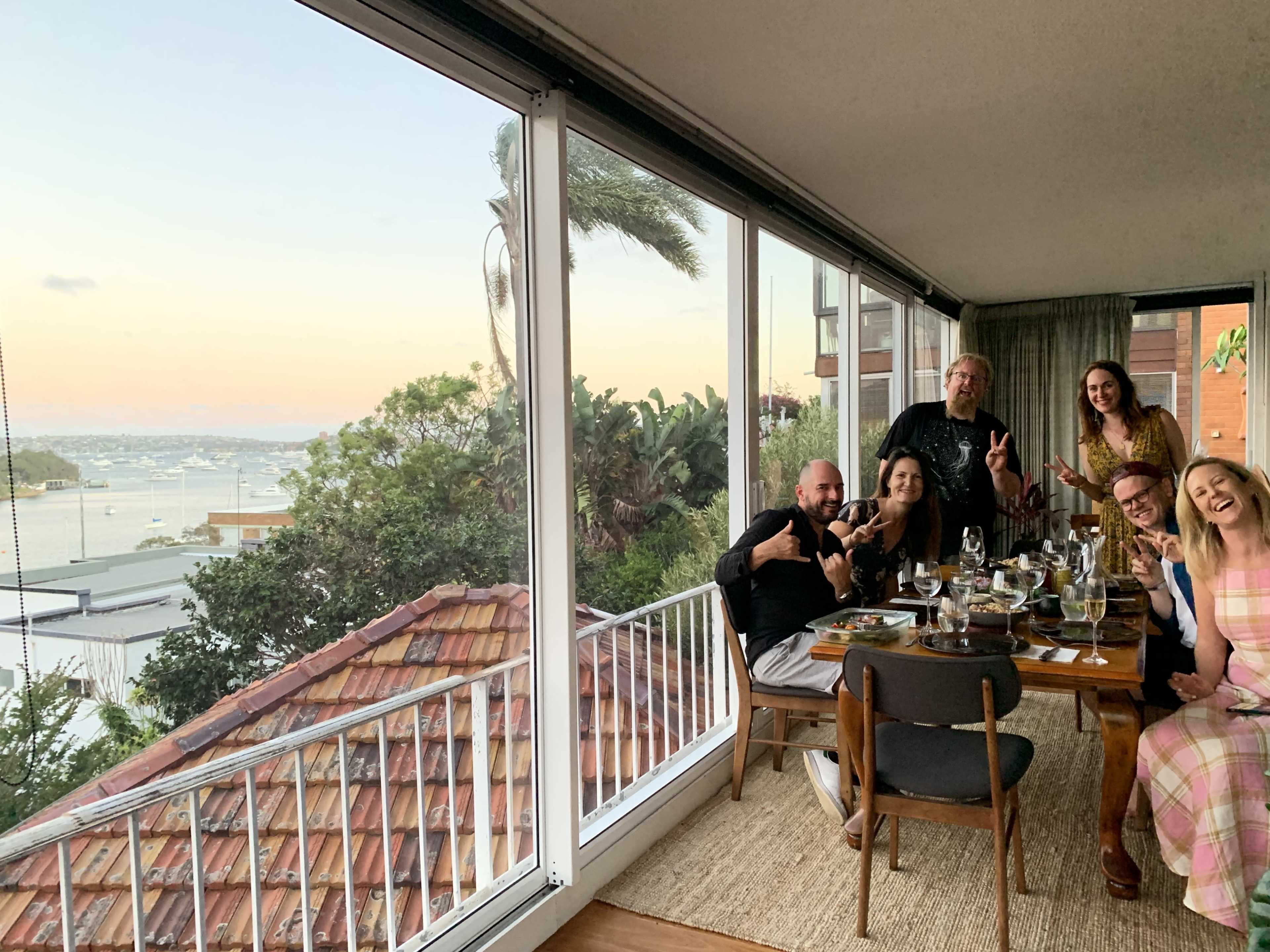 A group of six people is sitting at a dining table near a large window, enjoying a meal with a view of a body of water and a sunset in the background.