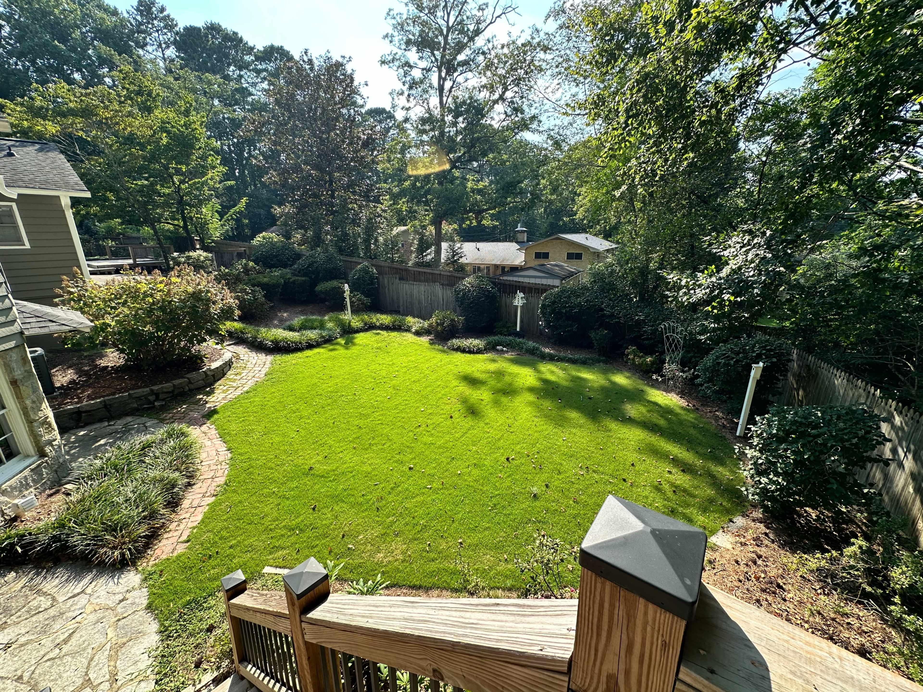 The image shows a well-maintained backyard with a green lawn, bordered by shrubs and trees, viewed from a wooden staircase.