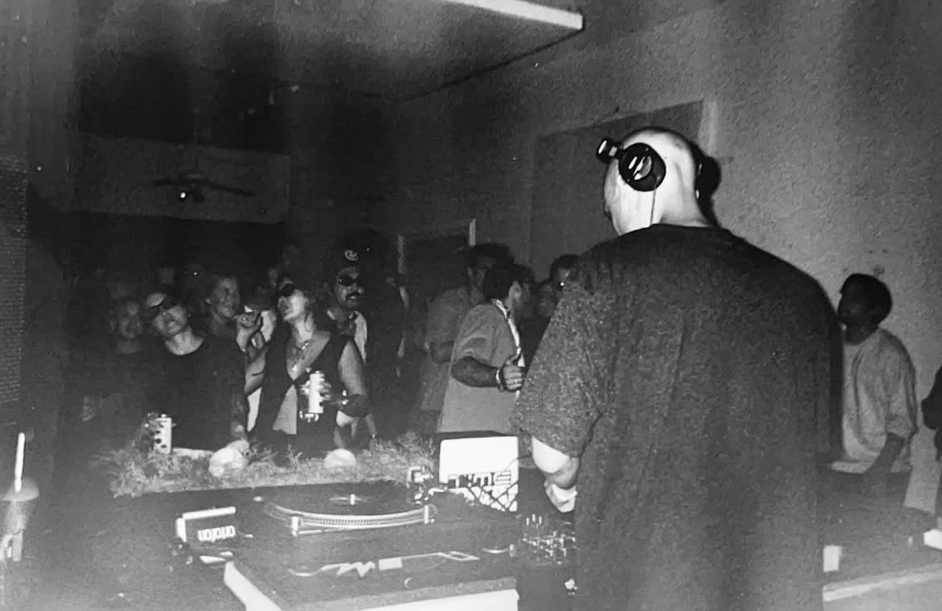 A DJ stands behind a turntable in a dimly lit room filled with people dancing and enjoying the music.